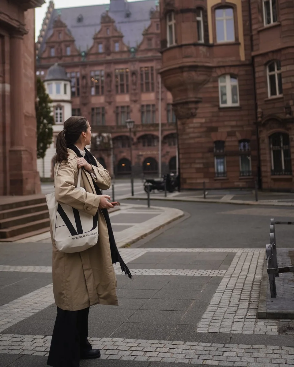 A woman standing on a city sidewalk in front of a historic red brick building with ornate architecture, holding a smartphone and wearing a trench coat and large tote bag.