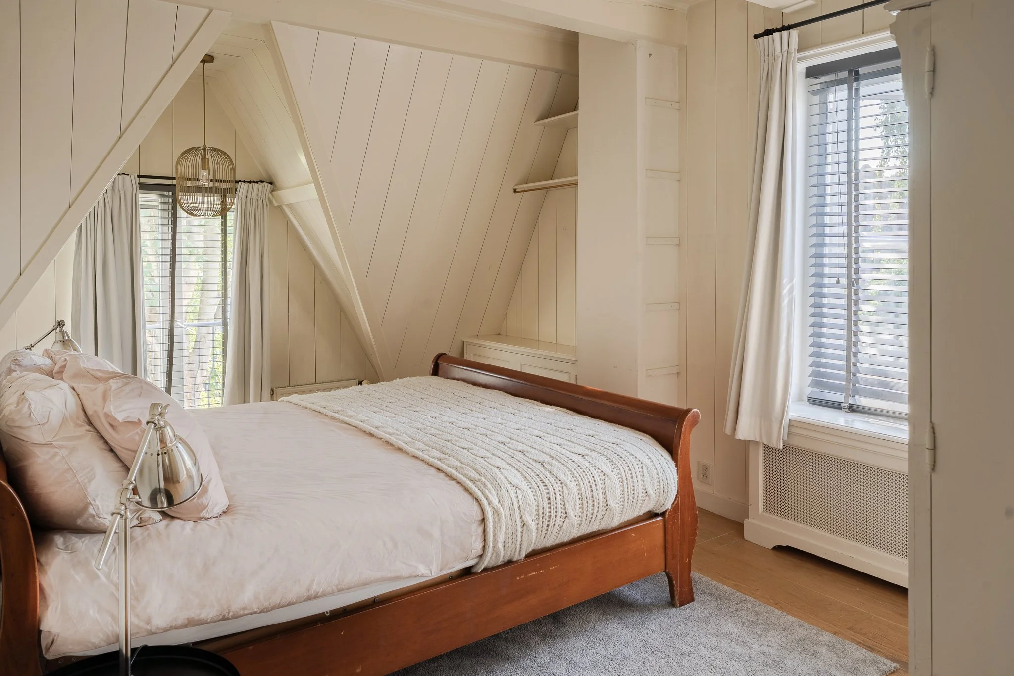A bedroom with a wooden bed, white bedding, and pink pillows. There are windows with white curtains on either side, and a ceiling light fixture. The walls are paneled in white.