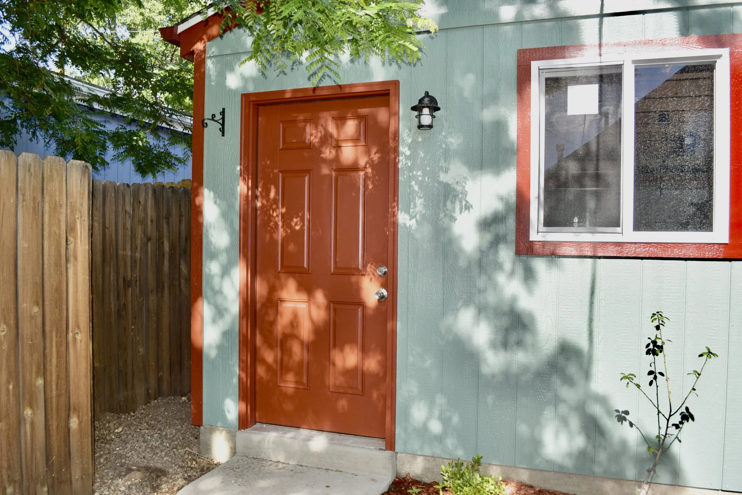 A red front door with a black outdoor wall lamp, set into a teal house with white trim around a window, next to a wooden fence and some plants, under a leafy tree.