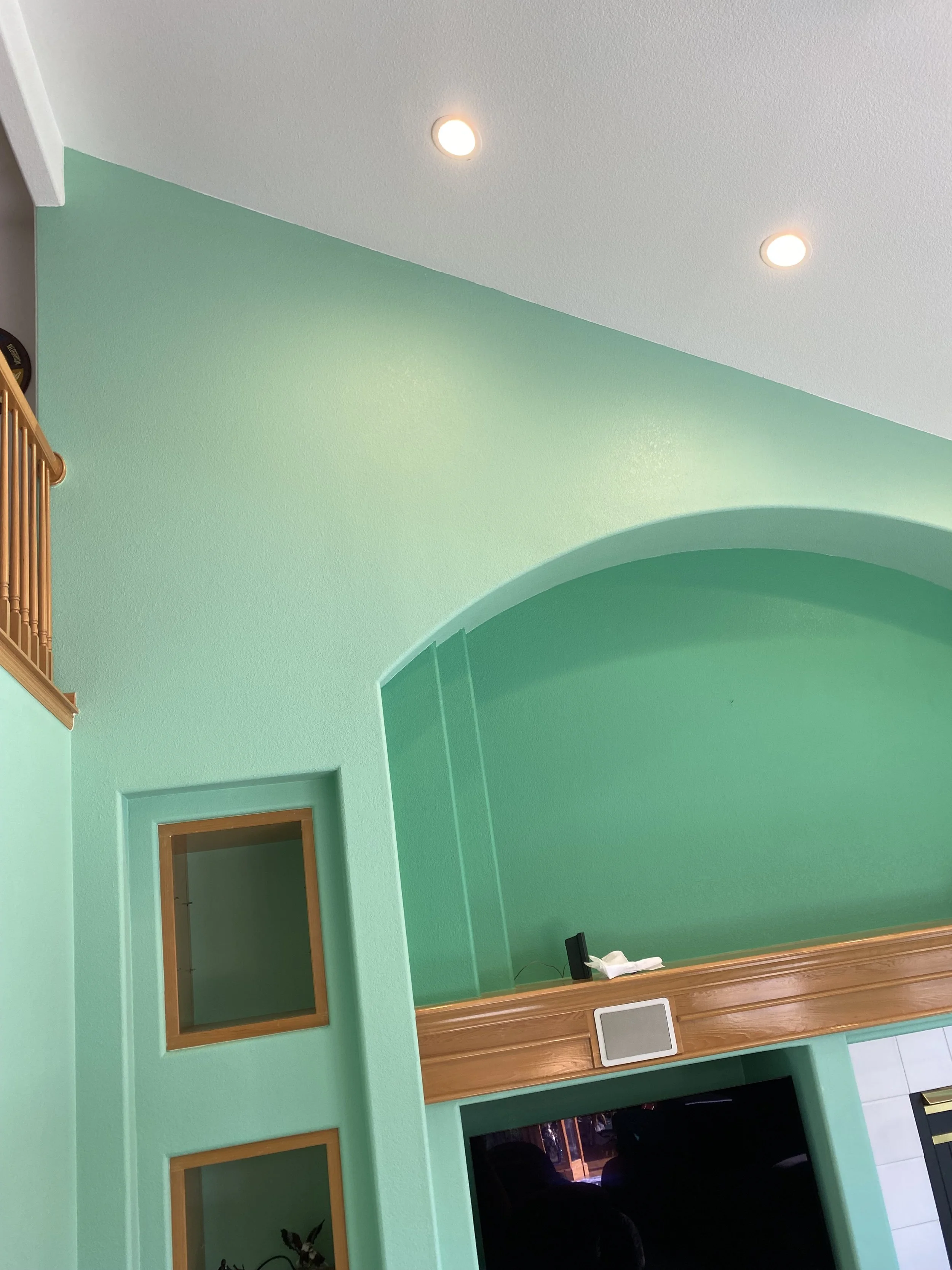 Indoor view of a room with mint green walls, two ceiling lights, and a wooden counter with a small gray speaker and white items on it. There are two wooden-framed mirrors and a door in the background.