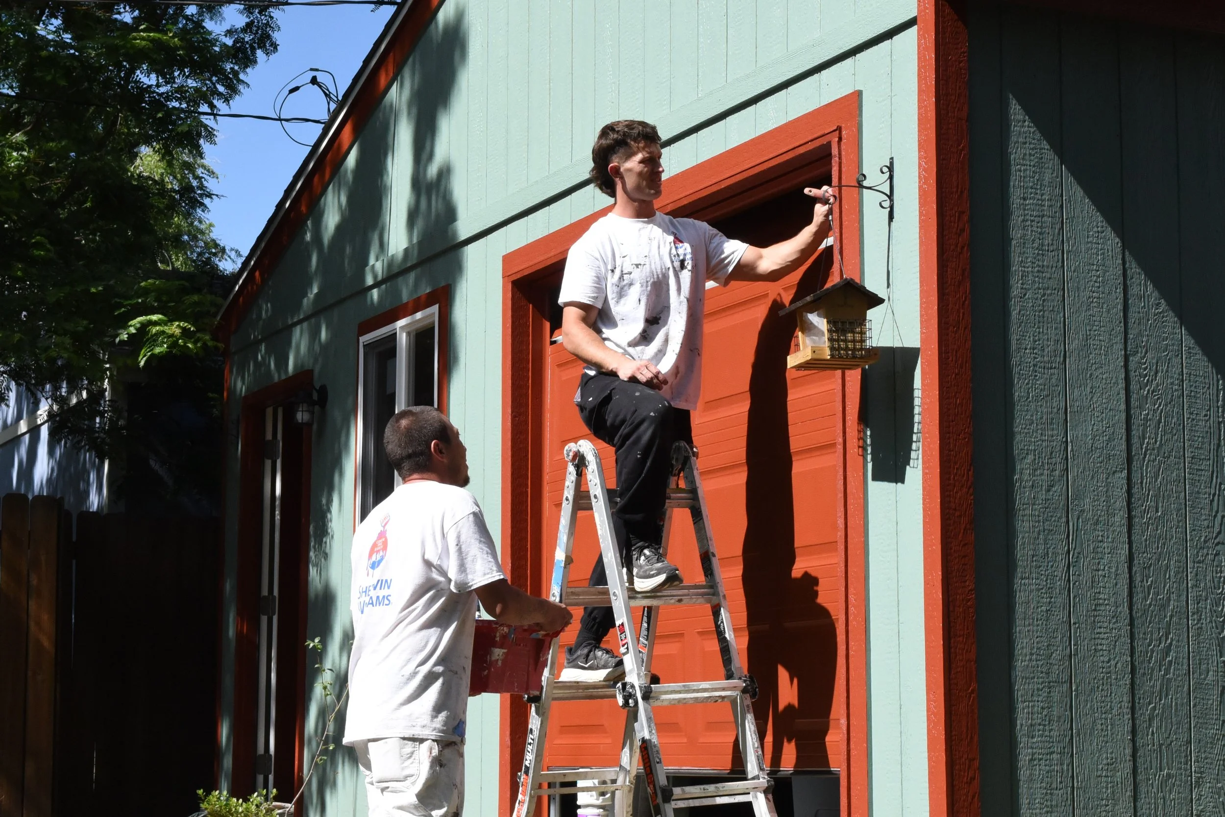 A man on a ladder painting the trim around a window of a house painted in mint green with orange trim. Two other people are nearby, one standing and one looking up, supervising or assisting.