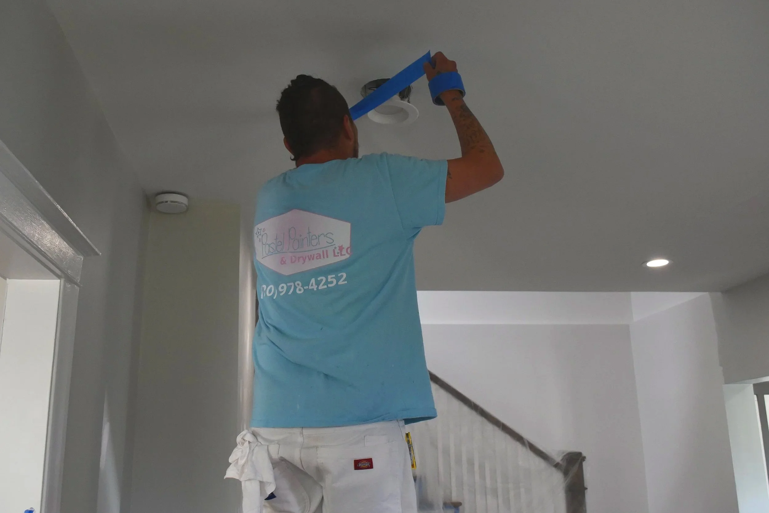 Man installing a ceiling light fixture using painter's tape on his hand, wearing a light blue t-shirt with business info, white pants, standing on a ladder in a house interior.