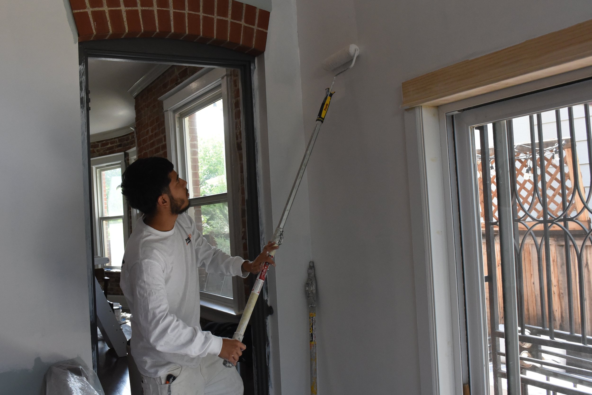 Person using a paint roller to paint the wall near a window in a room under renovation.