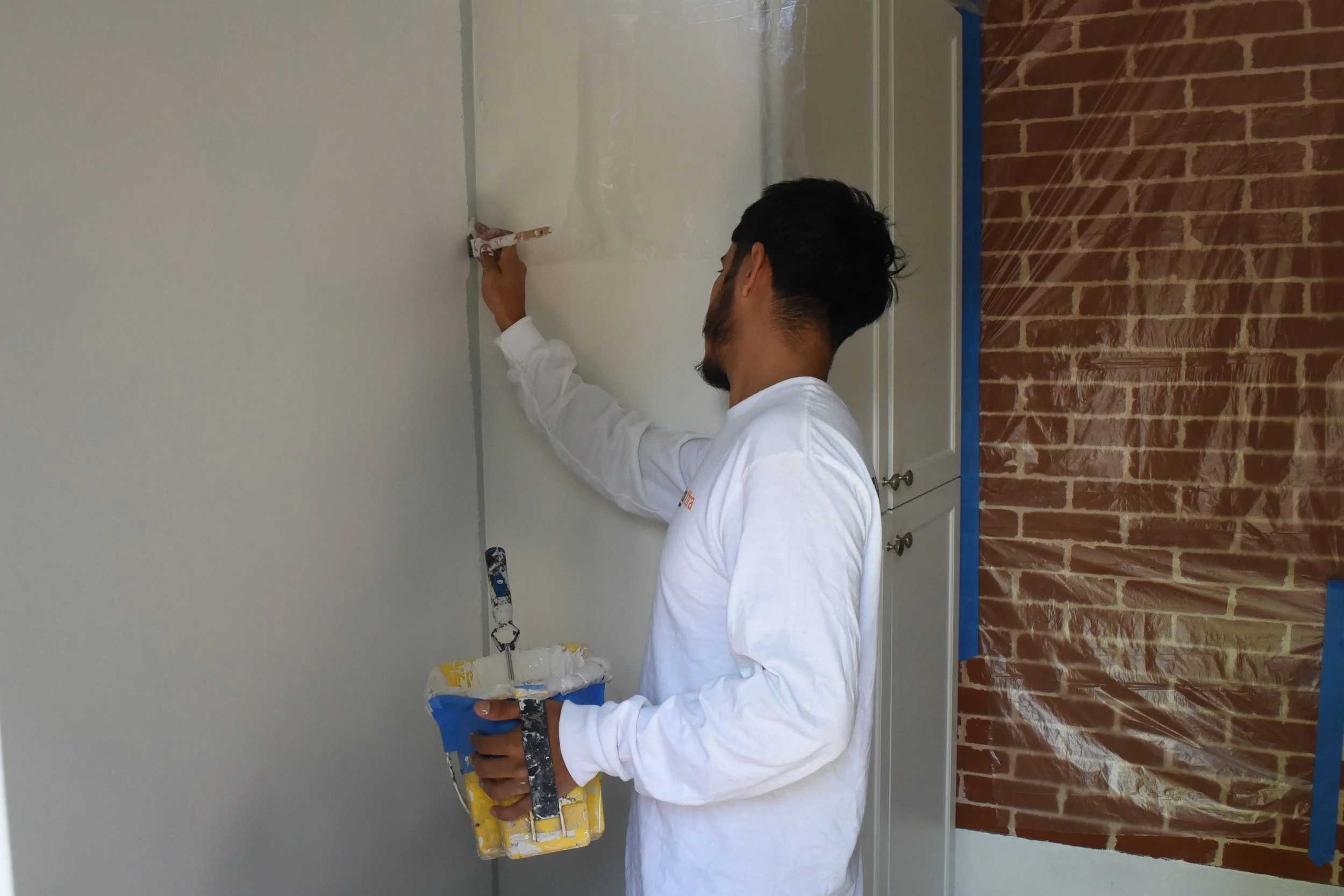 A man in a white long-sleeve shirt is painting a wall using a paint roller with a yellow paint tray nearby. The background includes a brick wall on the right side and a door with a lock in the middle of the wall.