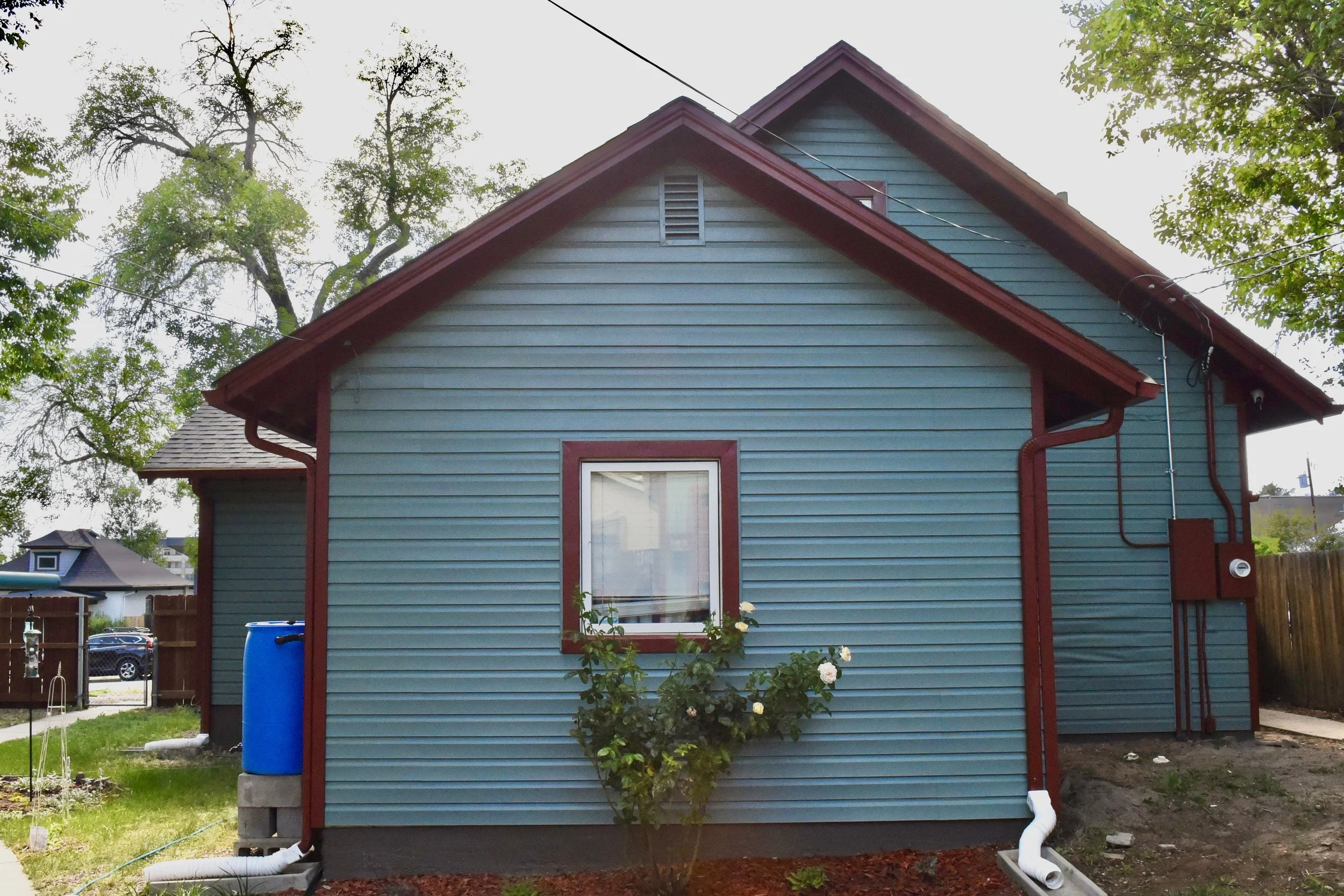 The image shows the side of a blue house with red trim, surrounded by trees and a grassy yard. A window with white curtains is visible in the lower part of the house. There are white and red gutters and downspouts, along with a blue rain barrel near the corner. A patio or yard with a fence and neighboring houses can be seen in the background.
