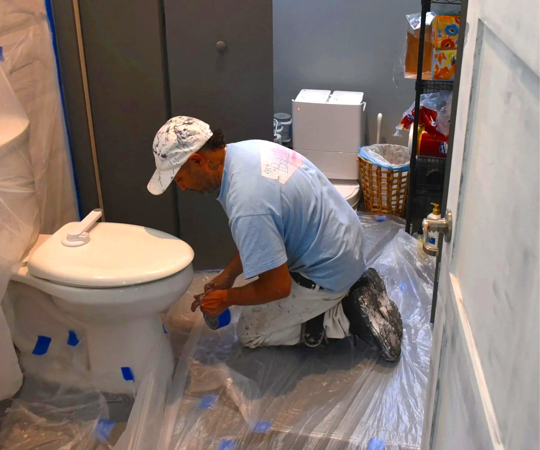 A person crouching on the floor working on a toilet, wearing a white cap and paint-stained clothing, surrounded by plastic sheets, in a room with various cleaning supplies and storage items.