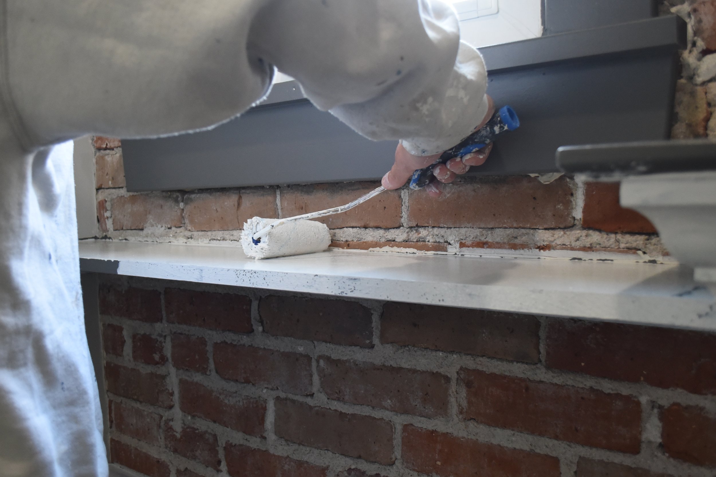 A person is using a paint roller to apply white paint to a brick wall. The person's hand and part of their arm are visible, and they are wearing a white long-sleeve shirt that is splattered with paint. The scene is indoors, with a focus on the painting process.