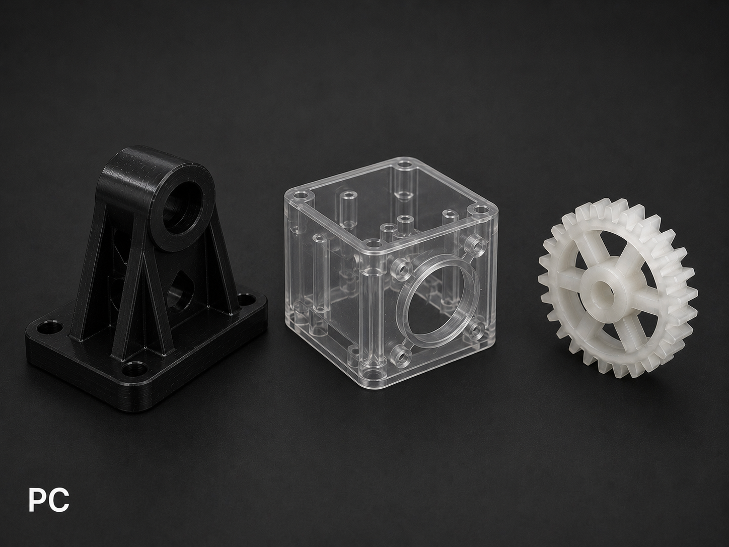 Three different plastic mechanical parts on a black background: a black L-shaped part with a cylindrical hole, a clear plastic cube with multiple holes, and a white gear with teeth.