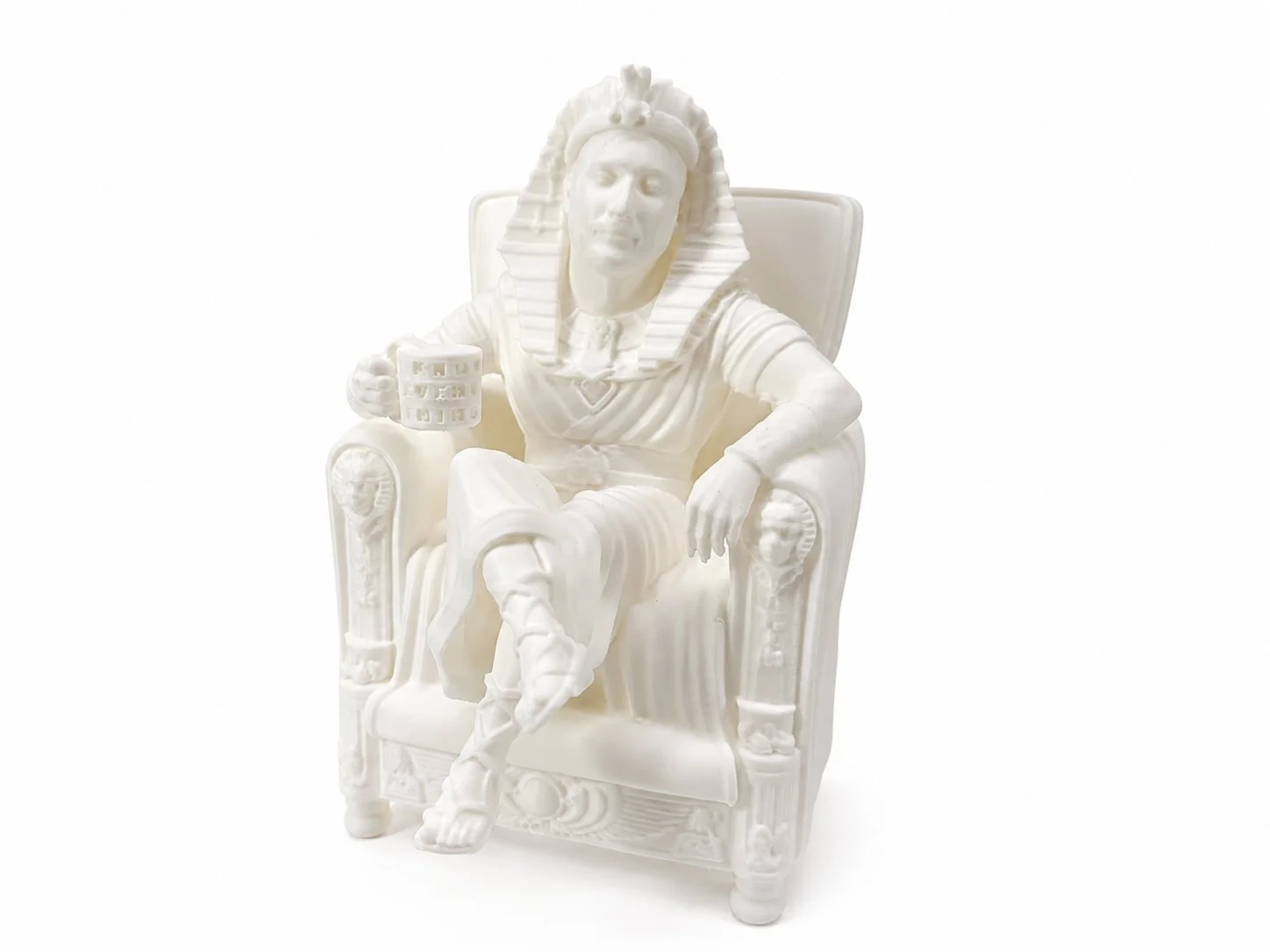 White sculpture of a person with a human face and a figure with an Egyptian headdress, sitting in a throne-like chair, holding a mug.