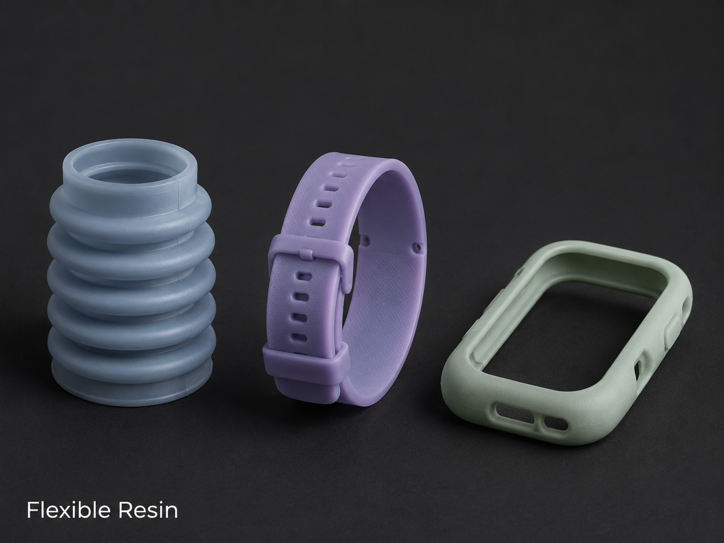 Three small, flexible resin objects in pastel colors: a blue coil, a purple watch strap, and a green watch frame, arranged on a dark surface.