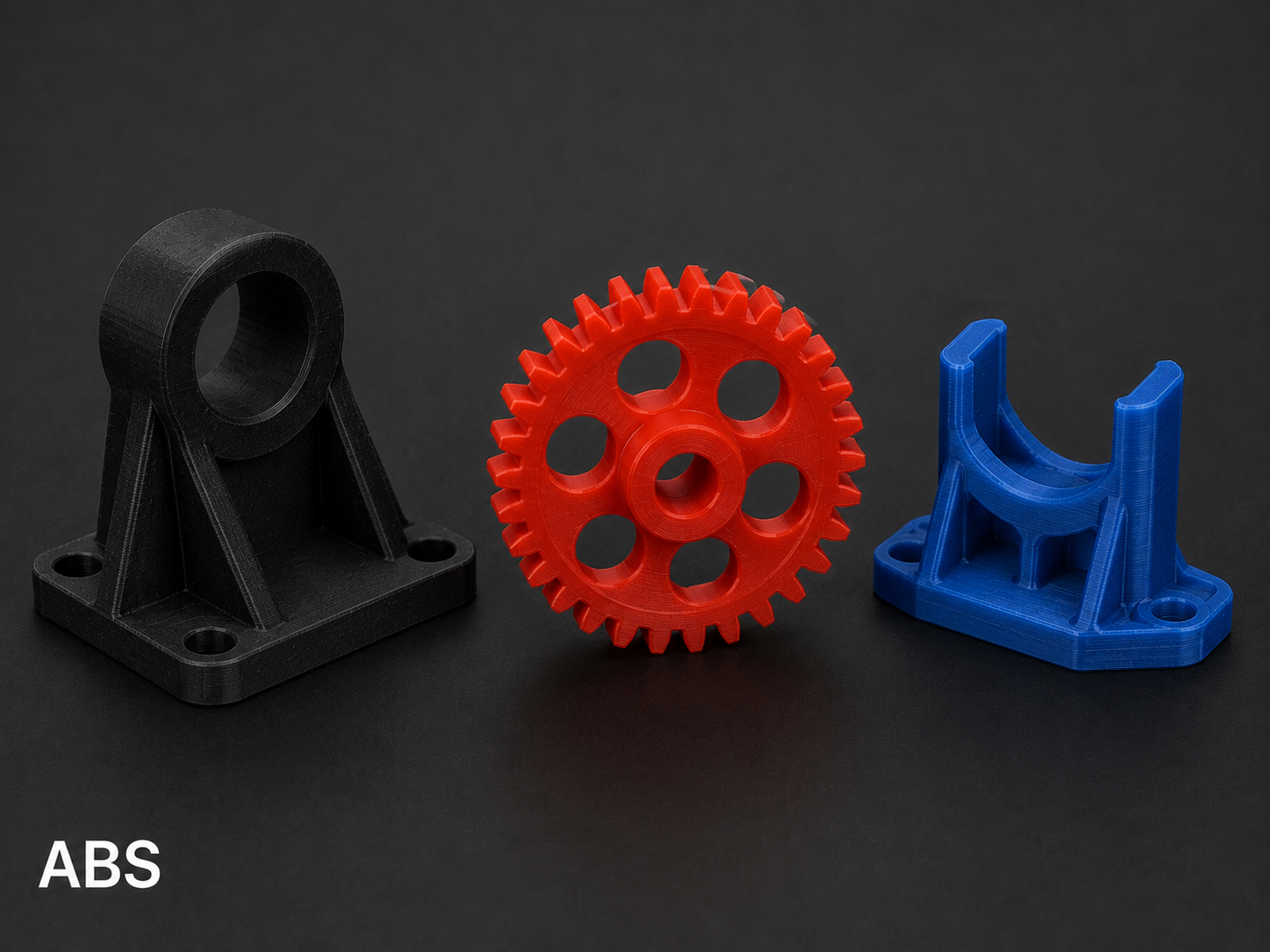 Three 3D-printed mechanical parts in red, black, and blue on black background, with the word 'ABS' in white at the bottom left corner.