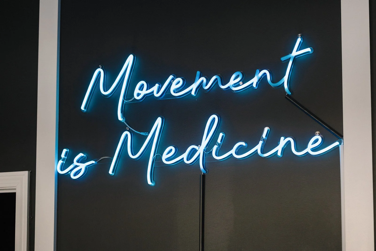 Blue neon sign that reads 'Movement is Medicine' on a dark wall implying movement is medicine for sobriety