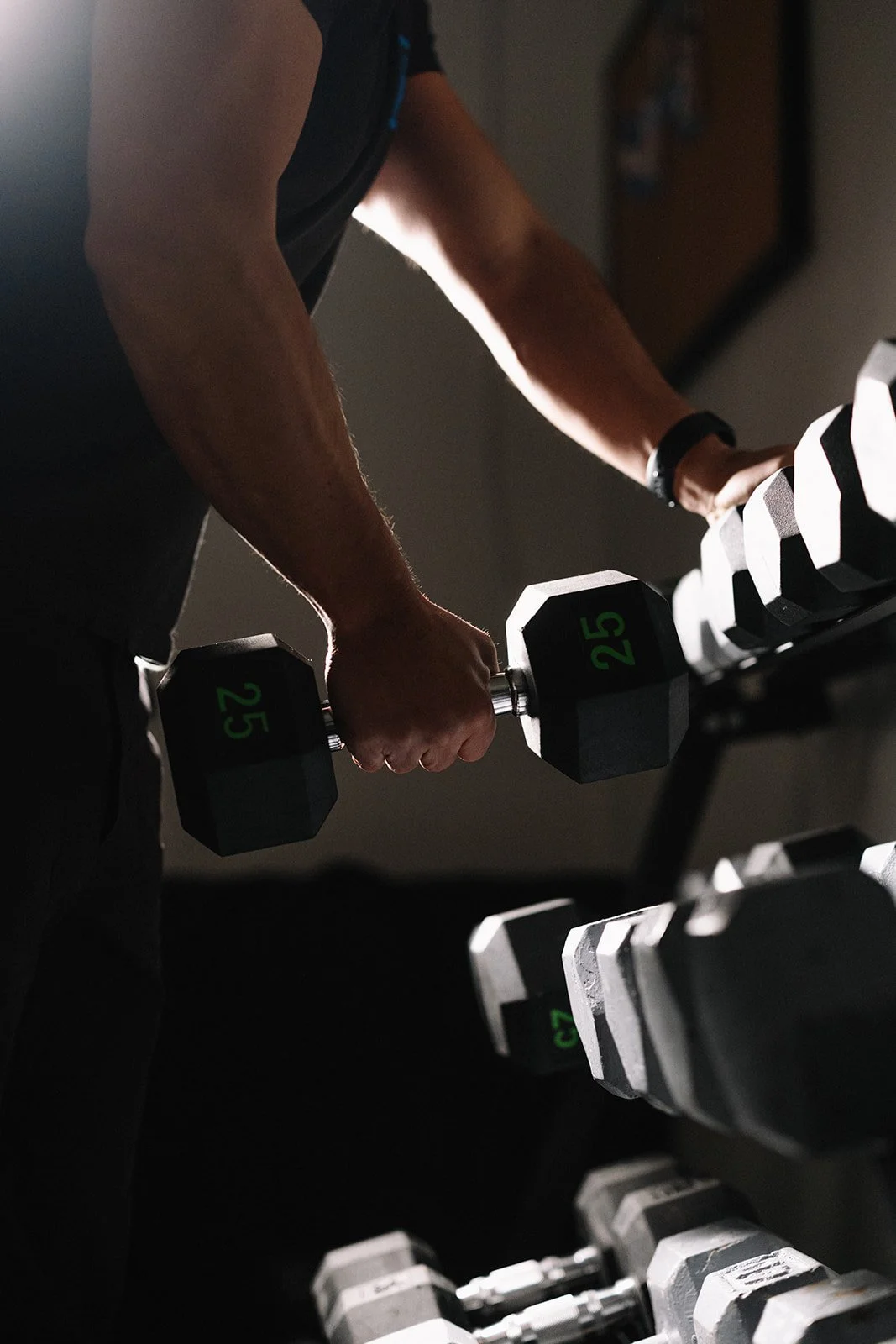 A sober person lifting a black dumbbell with green numbers in a gym, with other dumbbells on a rack beside them.