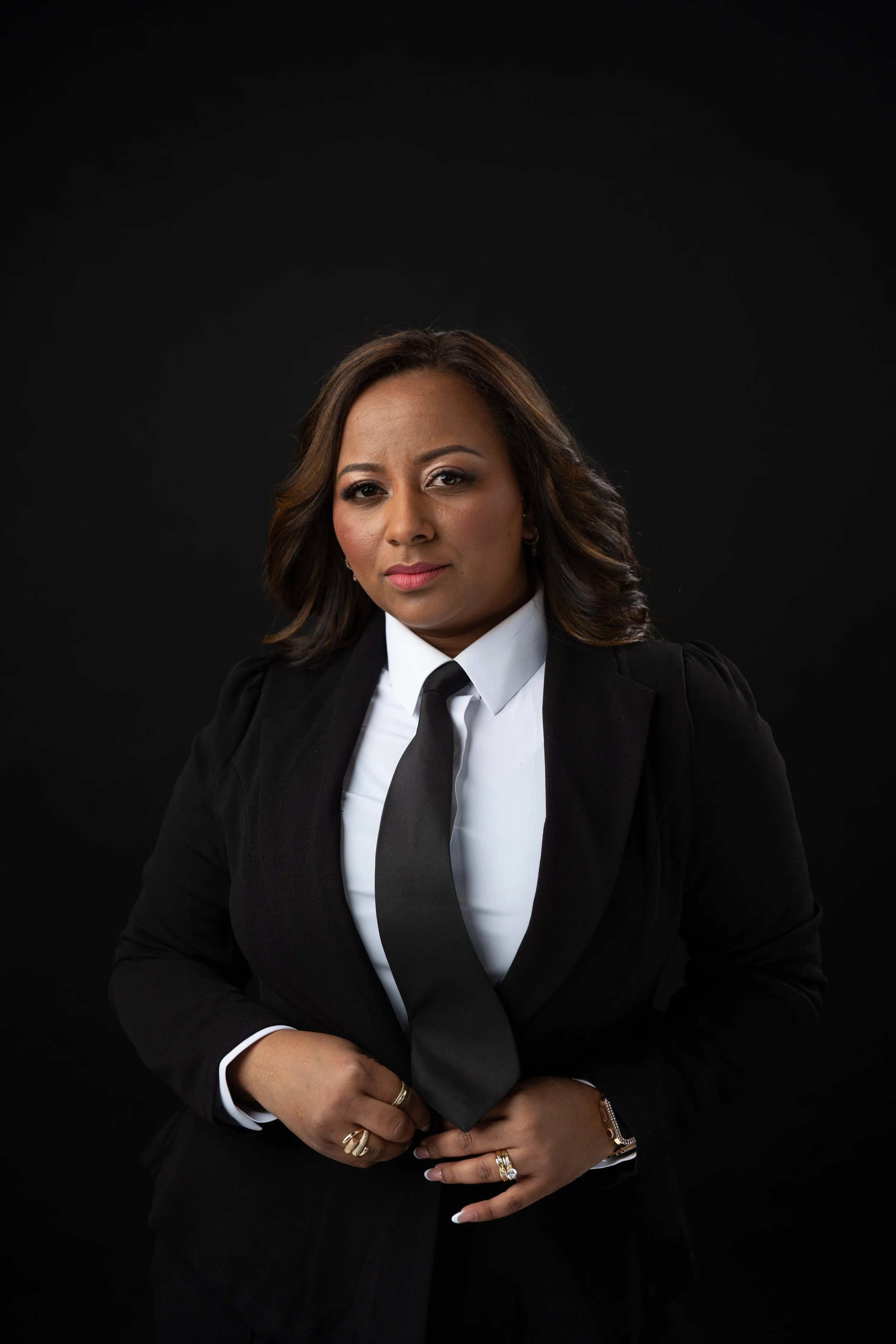 A confident woman in a black suit and white shirt, wearing a black tie, standing against a black background.