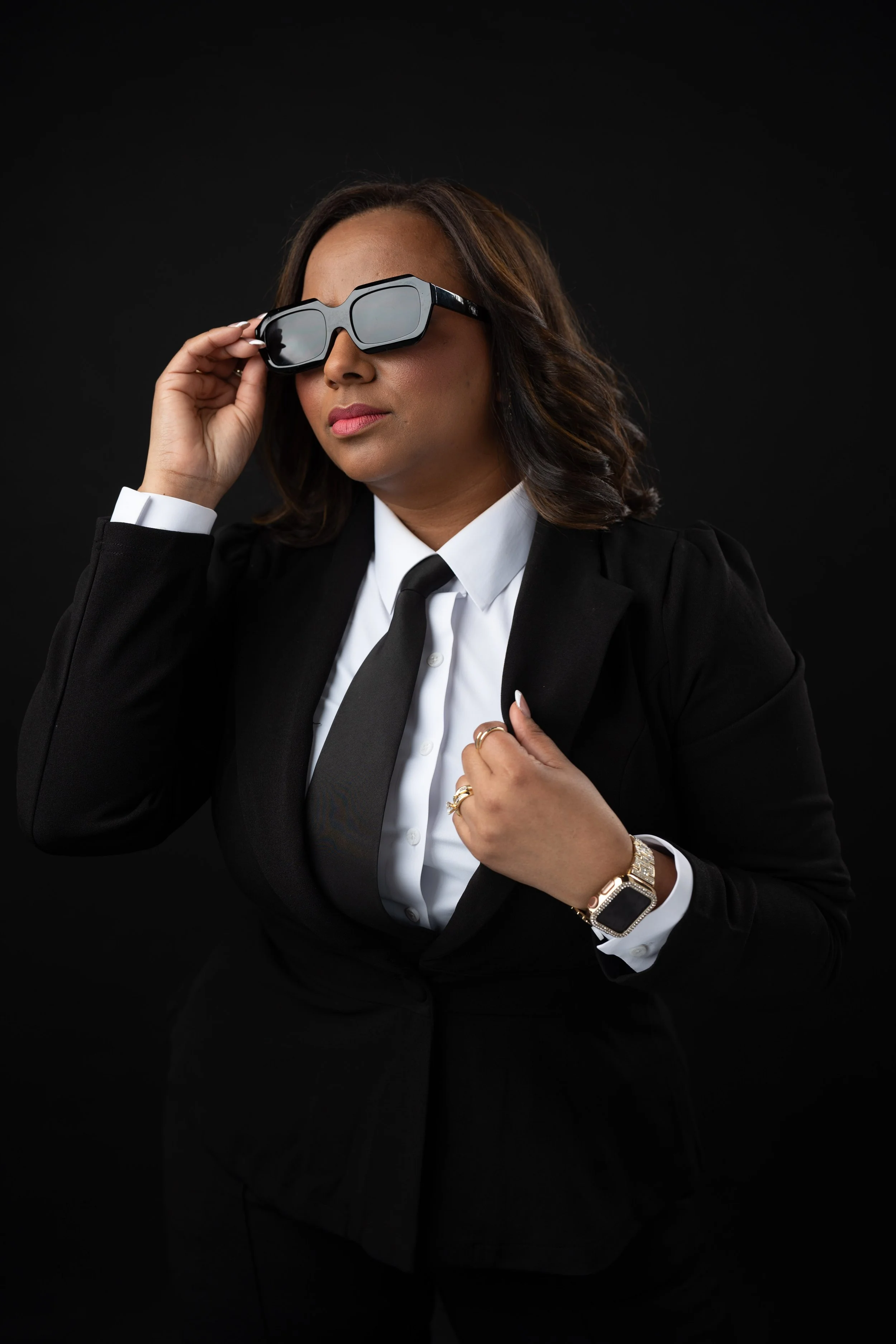 A confident woman dressed in a black suit and white shirt, wearing sunglasses, a watch, multiple rings, and adjusting her sunglasses against a black background.
