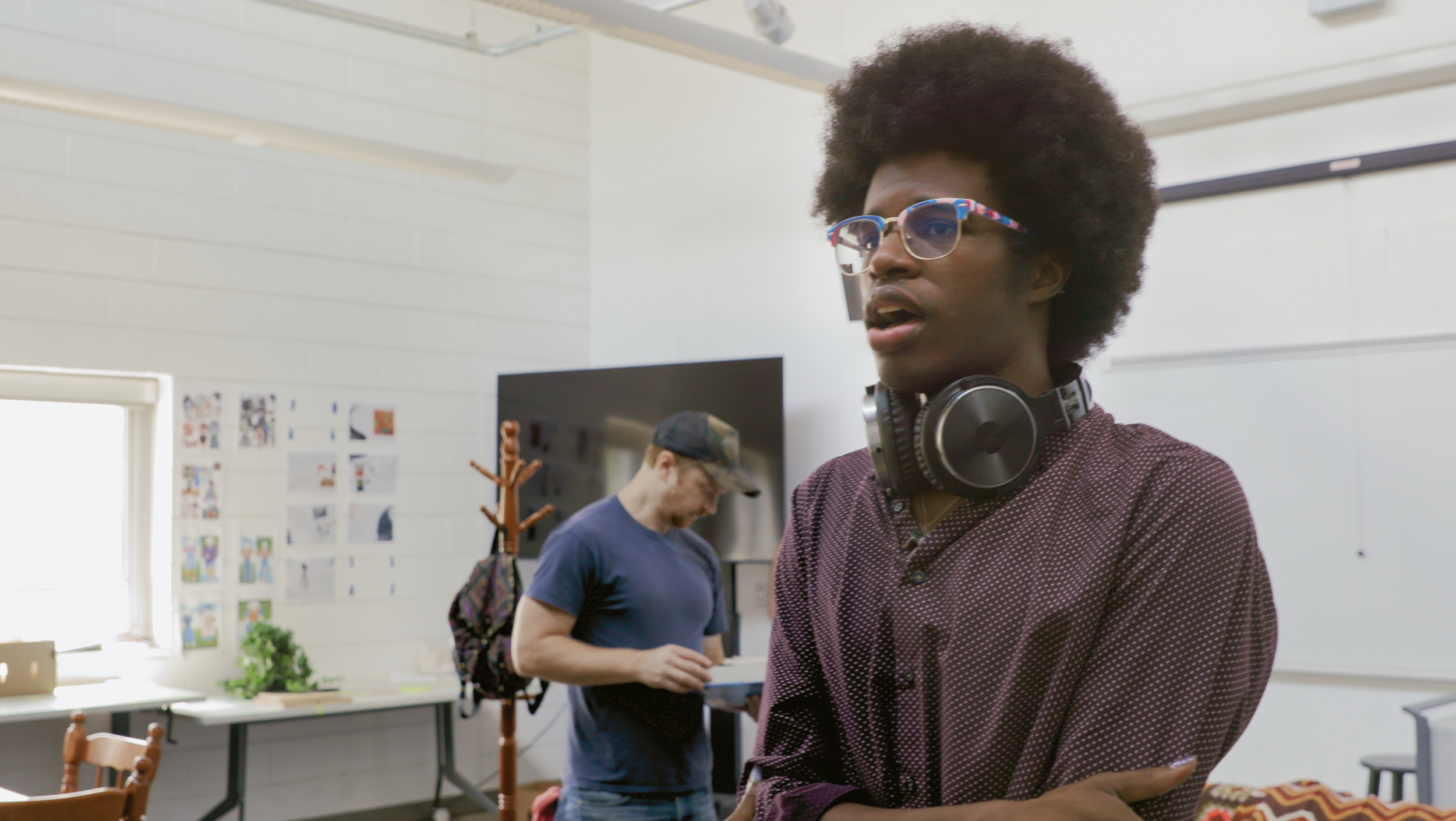 A person with curly hair and sunglasses, wearing headphones around their neck, stands with arms crossed in a classroom or office. In the background, another person with a cap is looking at a book, near a wall decorated with photos or posters.