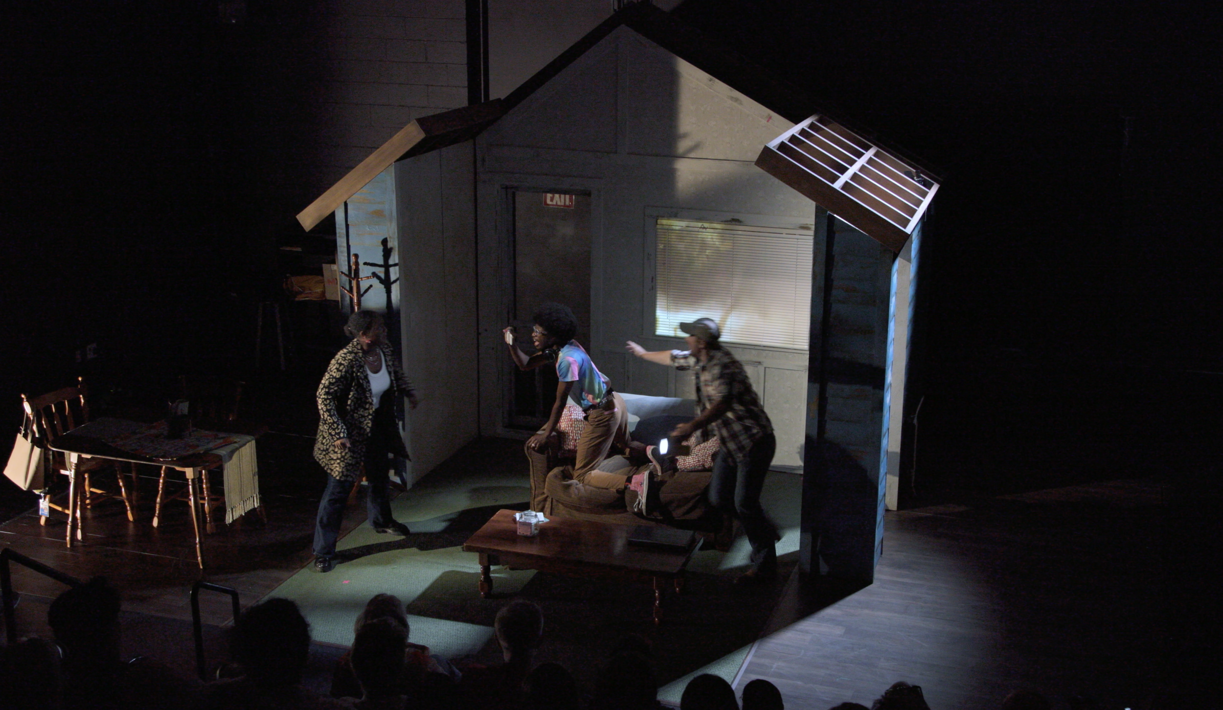 People acting on a small stage resembling a living room in a theater, with three individuals engaged in a scene, a table, and a chair set with a tablecloth, facing an audience.