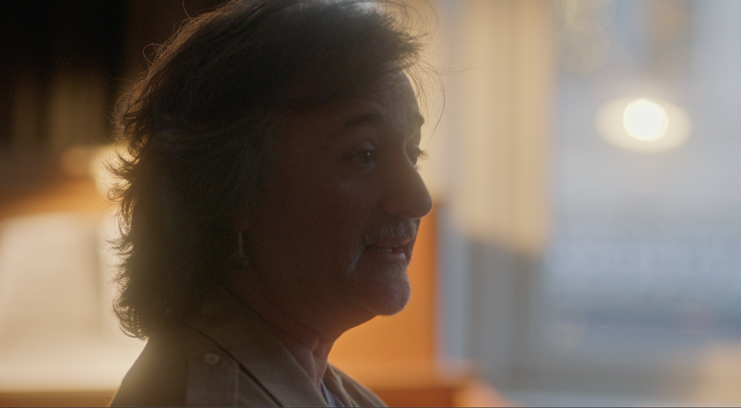 A person in profile with long, dark hair, in a warmly lit indoor setting.