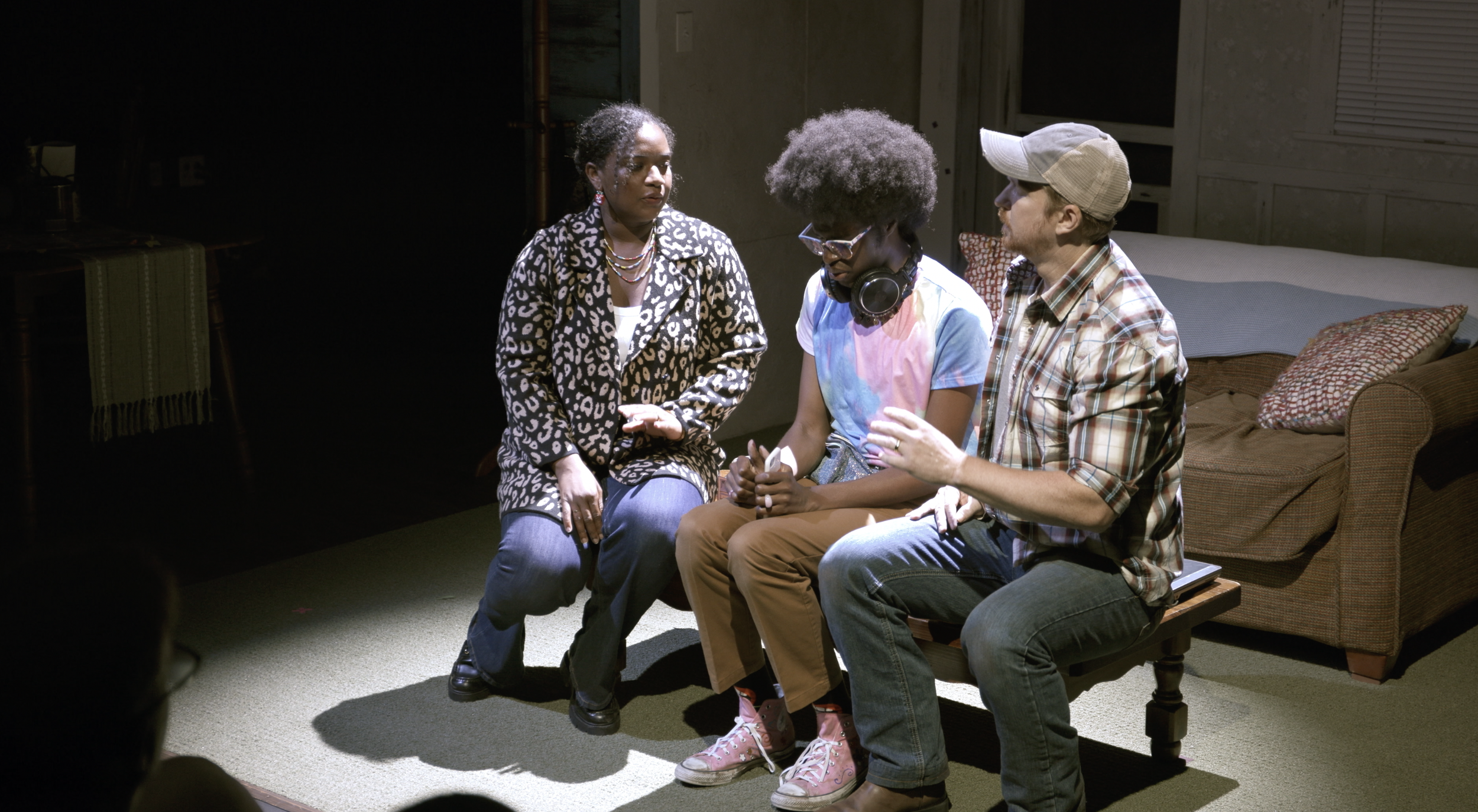 Three people sitting and talking in a dimly lit room, all appearing to be in a theatrical performance or rehearsal.
