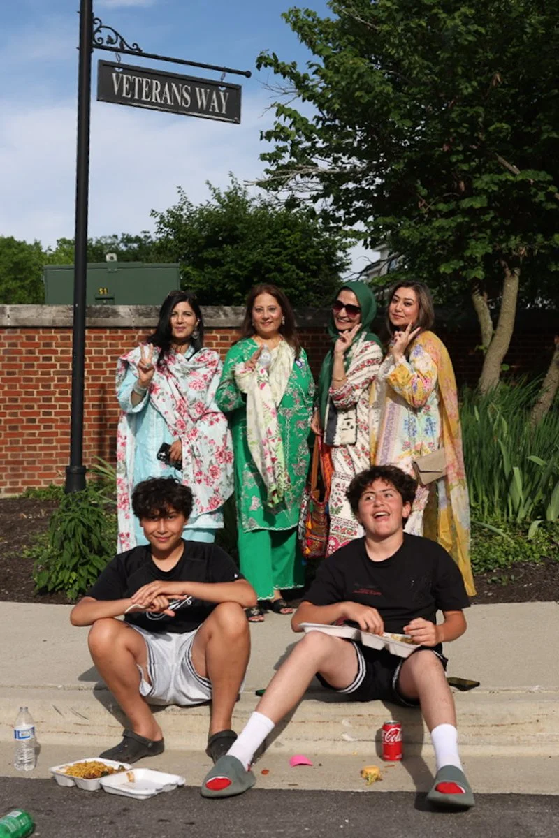 A group of people in traditional South Asian clothing standing and sitting near a Veterans Way sign during the Carmel Eid Festival.