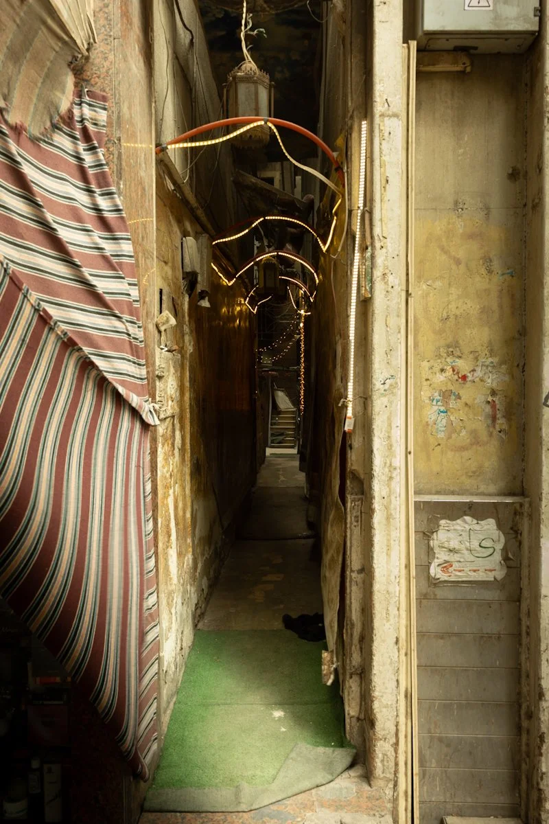Dim narrow alley with peeling walls, striped fabric, and lights leading to staircase