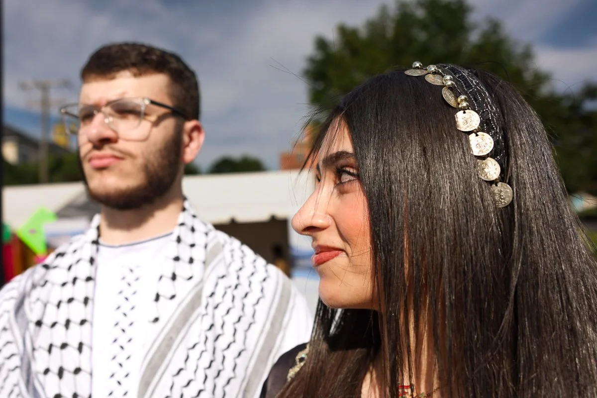 A brother and sister standing together at an outdoor festival with tents and trees in the background.