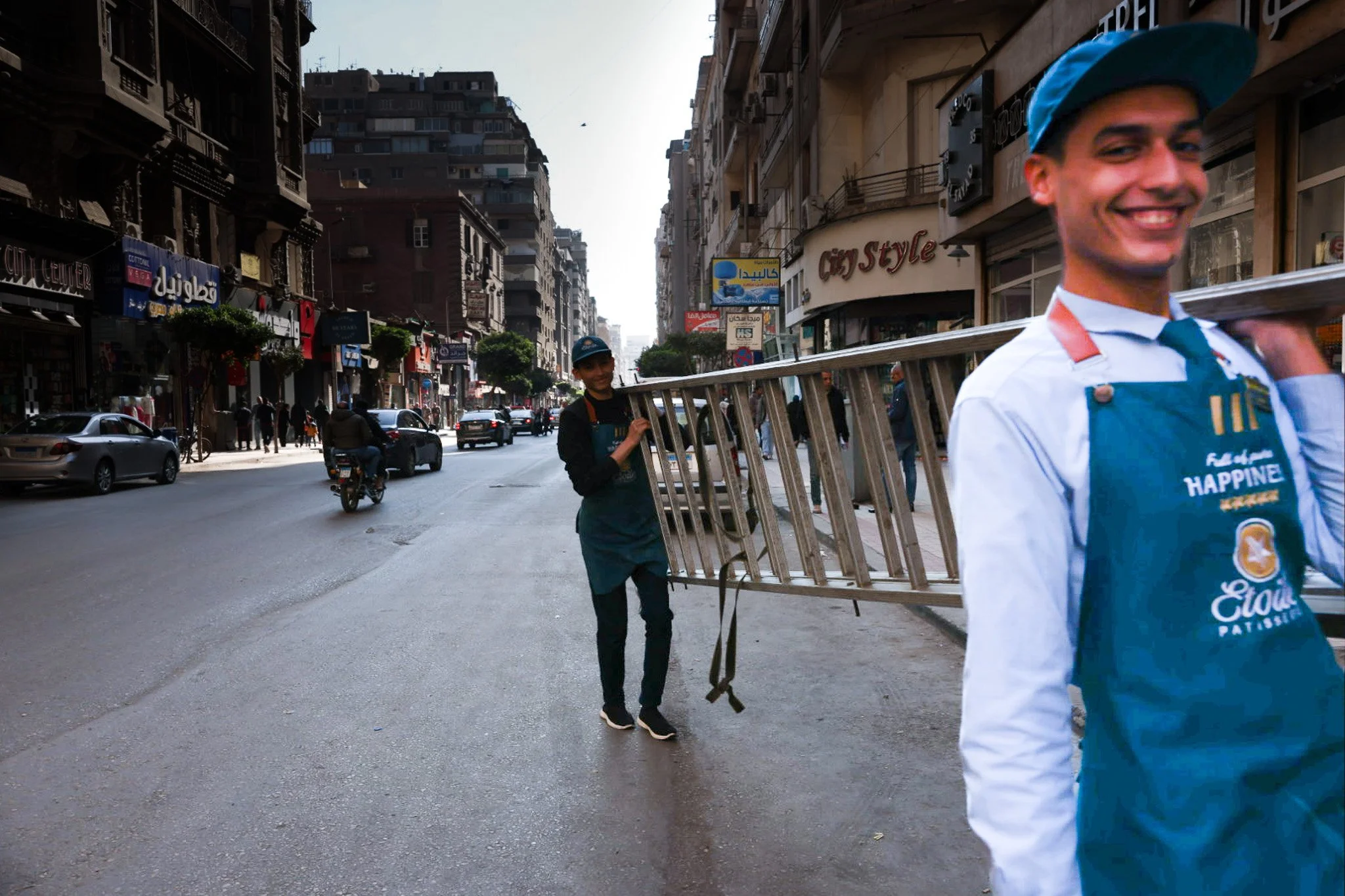 Two workers in blue aprons carrying ladder across urban street