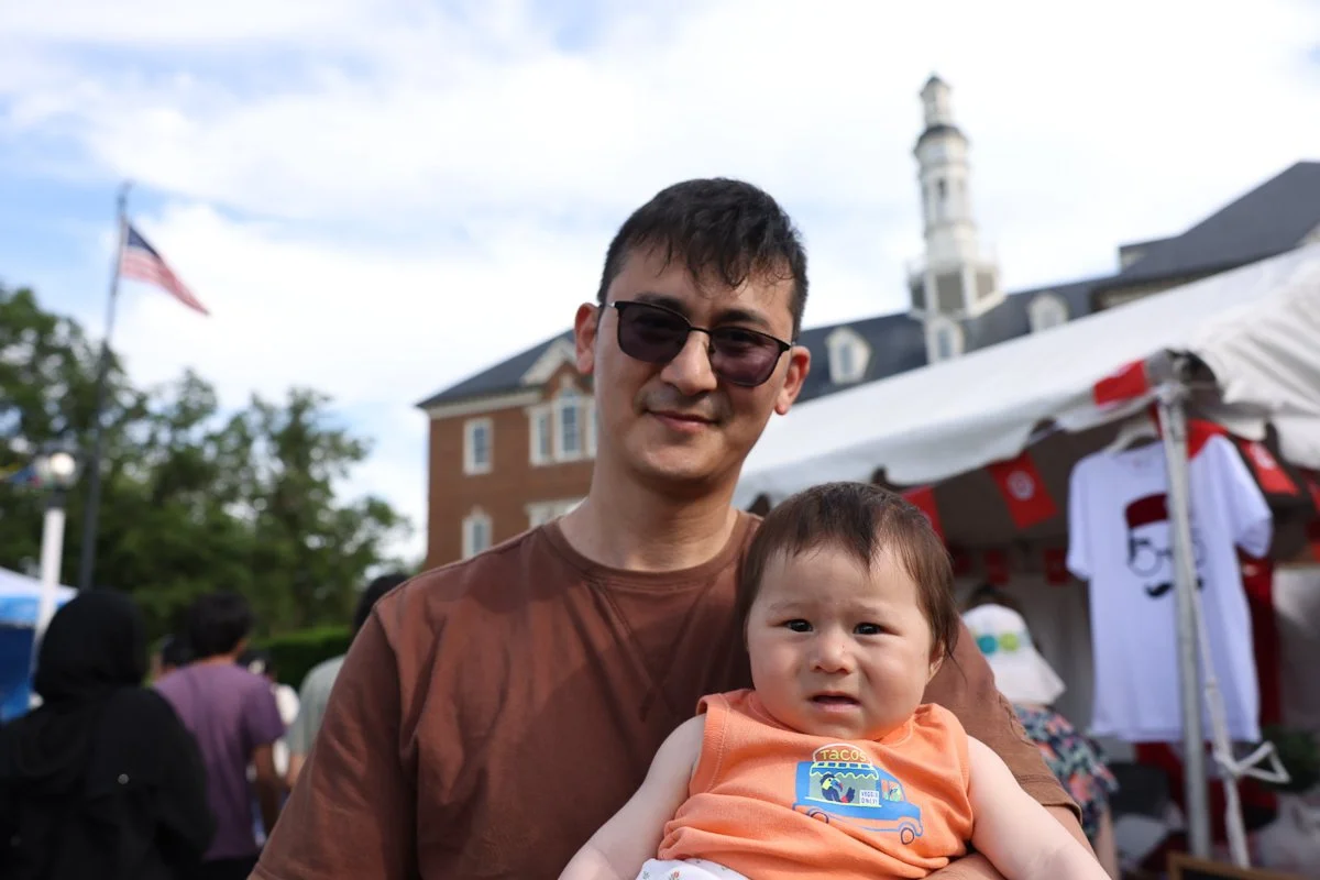 A man from Uzbekistan holding a baby at an outdoor festival with tents and people in the background.