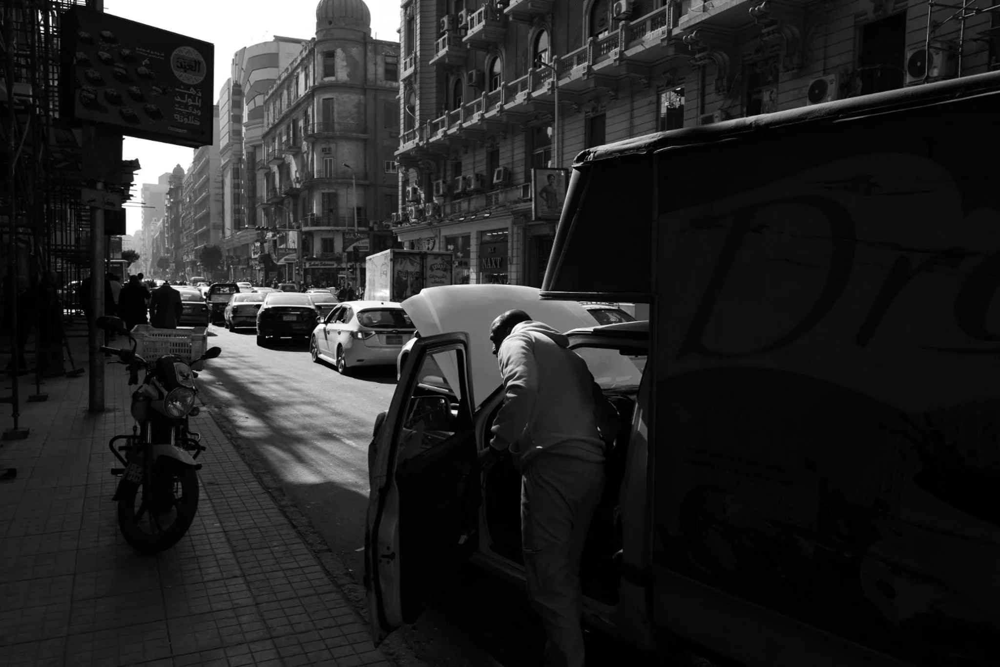 Person leaning into open‑hood car on busy street with ornate buildings