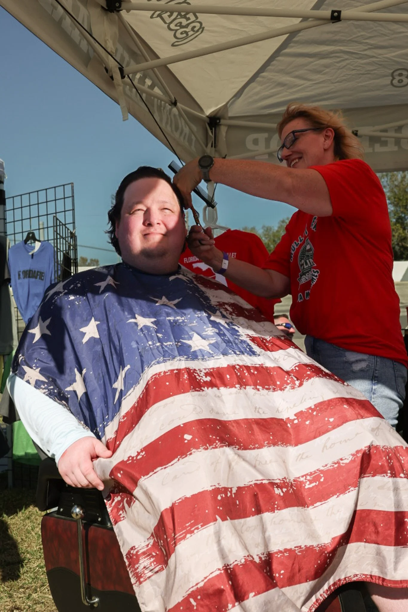 A man sitting in a wheelchair getting a shave outdoors, covered with a blanket featuring the American flag, under a canopy tent with a woman in a red shirt providing the service.