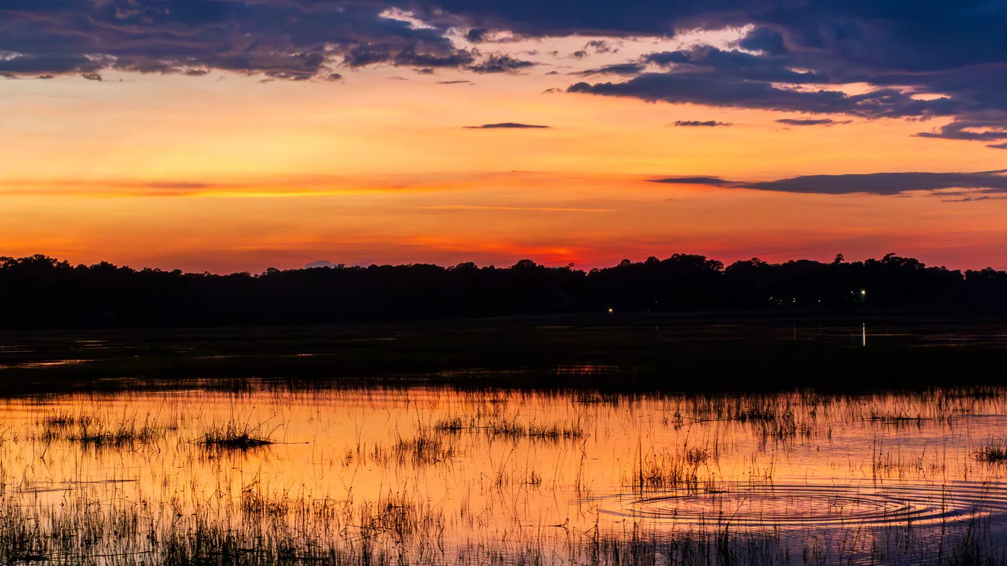 A scenic sunset over a body of water with reflections, silhouetted trees in the distance, and colorful clouds in the sky.