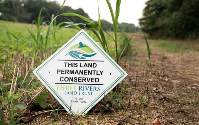 A sign planted in the ground that reads, "This land permanently conserved" by Three Rivers Land Trust, with a logo of a river, grass, and hills, in a rural area with soil, grass, and trees in the background.