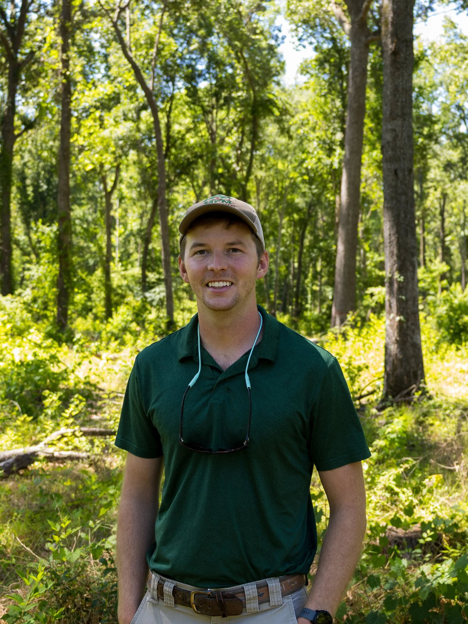 Young man smiling outdoors in a green shirt, beige pants, a tan cap, in a wooded area with tall trees and green foliage.