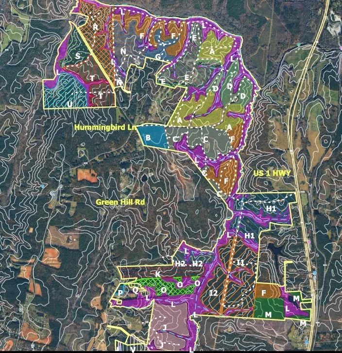 A detailed map of a hilly area showing various zones, roads, and trails with labels. Major roads include US Highway 1 and Green Hill Road. The map features different colored regions with distinct boundaries and labels, including areas labeled A through M, and specific trails such as Hummingbird Lake and multiple other trails marked with H, I, J, K, and L. Topography is indicated by contour lines, and some areas are shaded in purple, brown, green, and other colors for different zones.