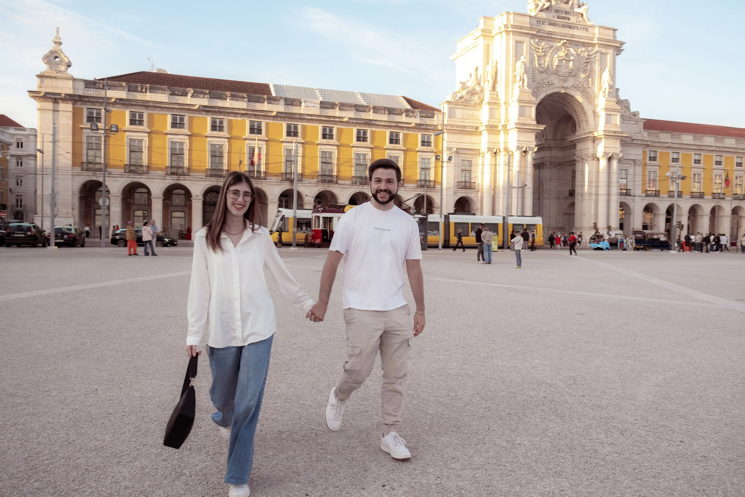 Casal andando de mãos dadas em uma praça na frente de um edifício histórico com arquitetura clássica e detalhes ornamentais, ao entardecer.