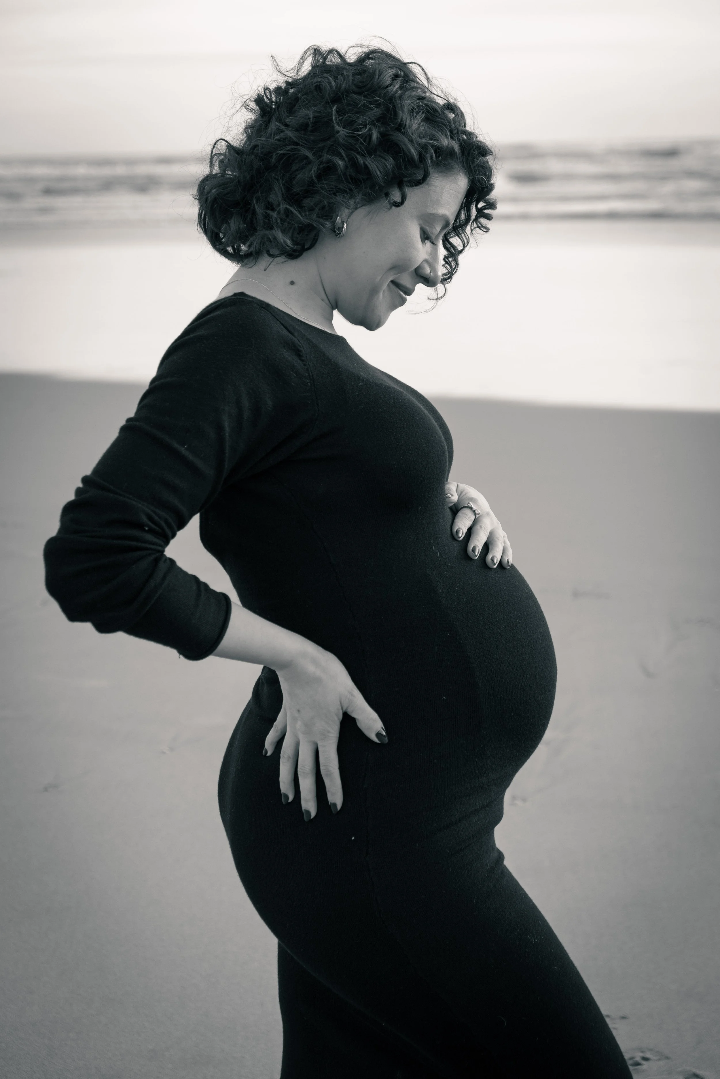 Retrato de uma mulher grávida com cabelo cacheado, de camiseta preta, com as mãos na barriga, na praia ao pôr do sol, em tom preto e branco.