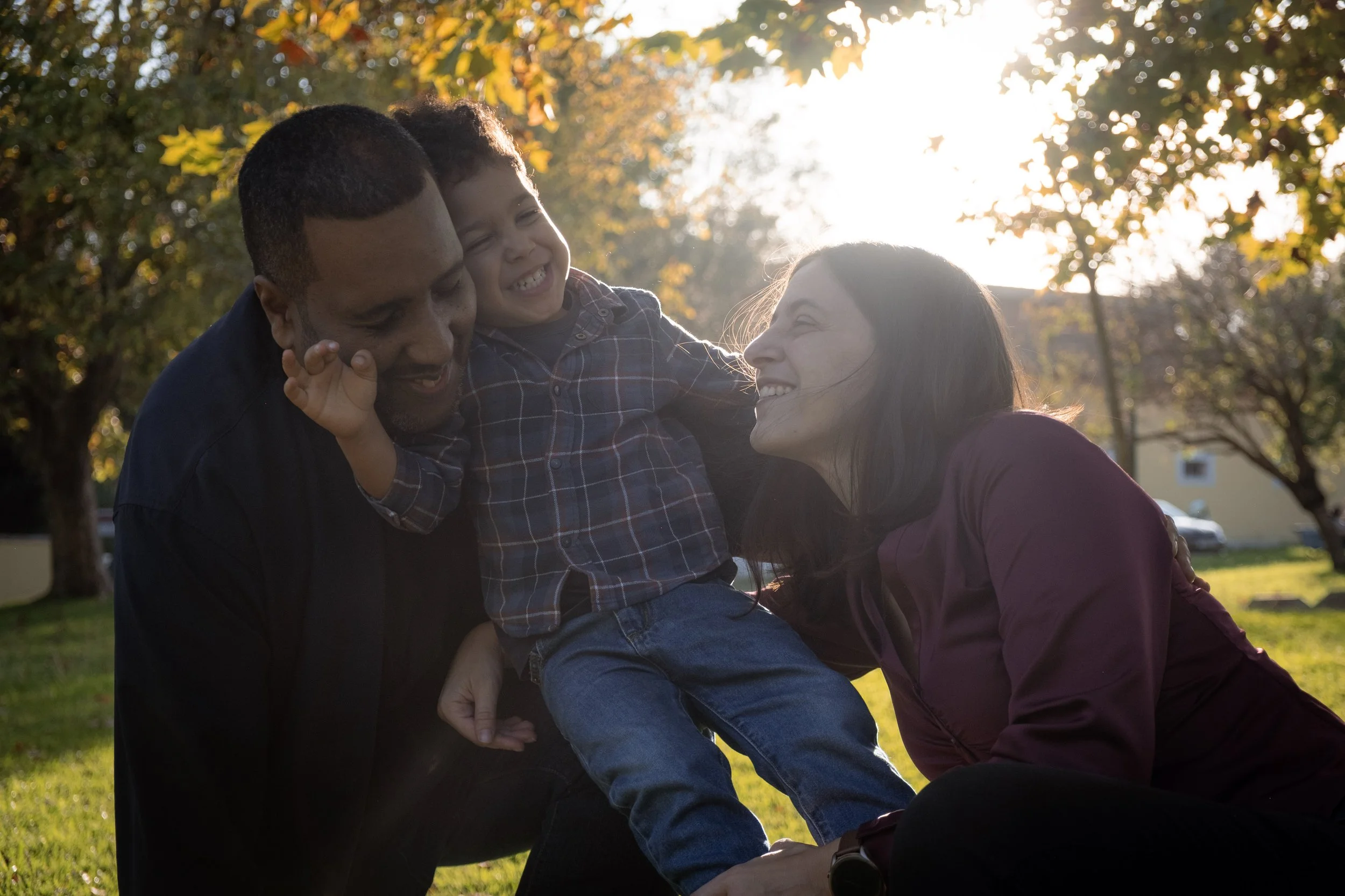 Família brincando e sorrindo ao ar livre, sob a luz do sol, com árvores ao fundo.