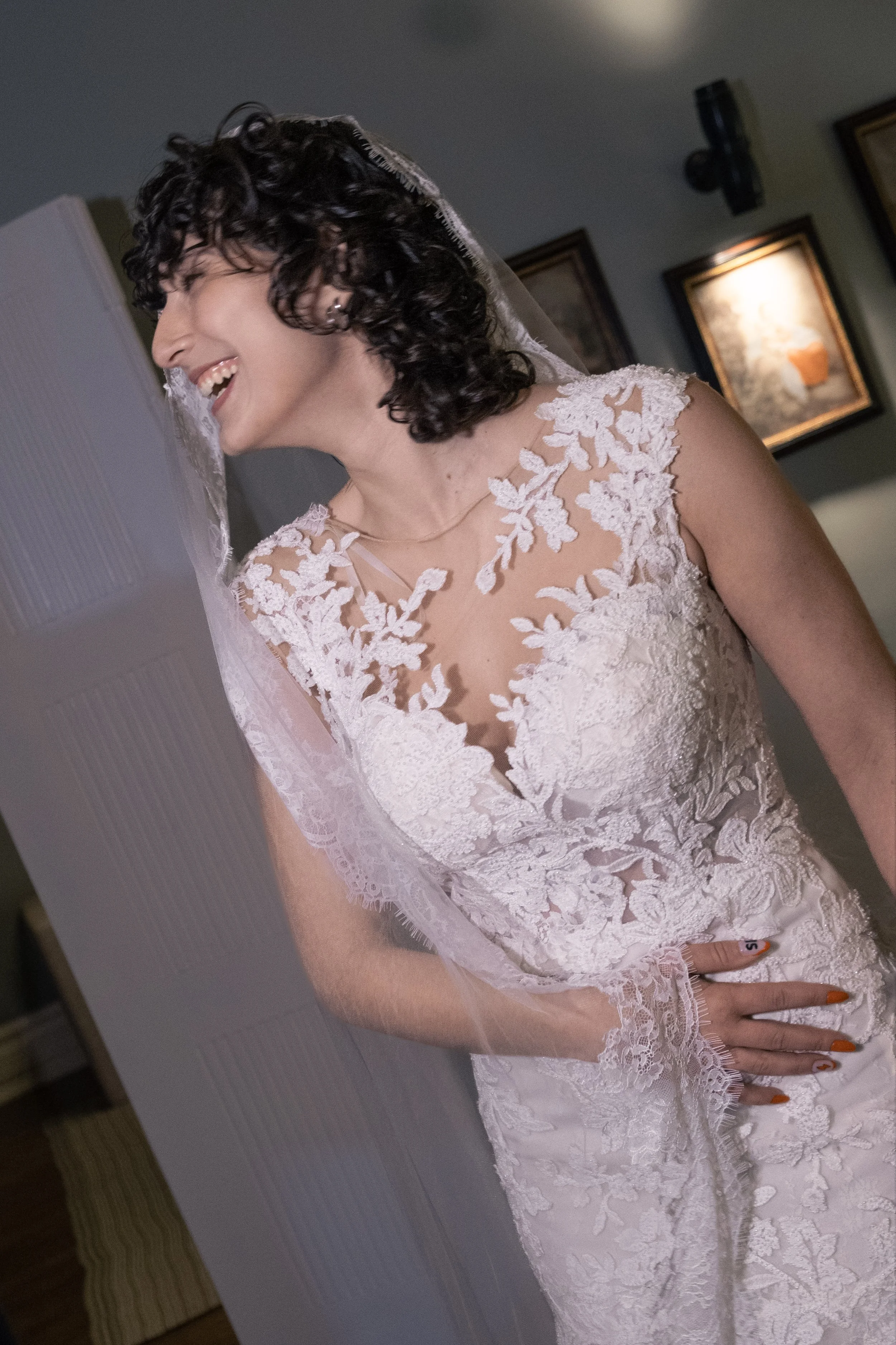 Mulher sorrindo vestida com vestido de noiva de renda com detalhes florais, usando luvas e véu, com esmaltes laranja na mão