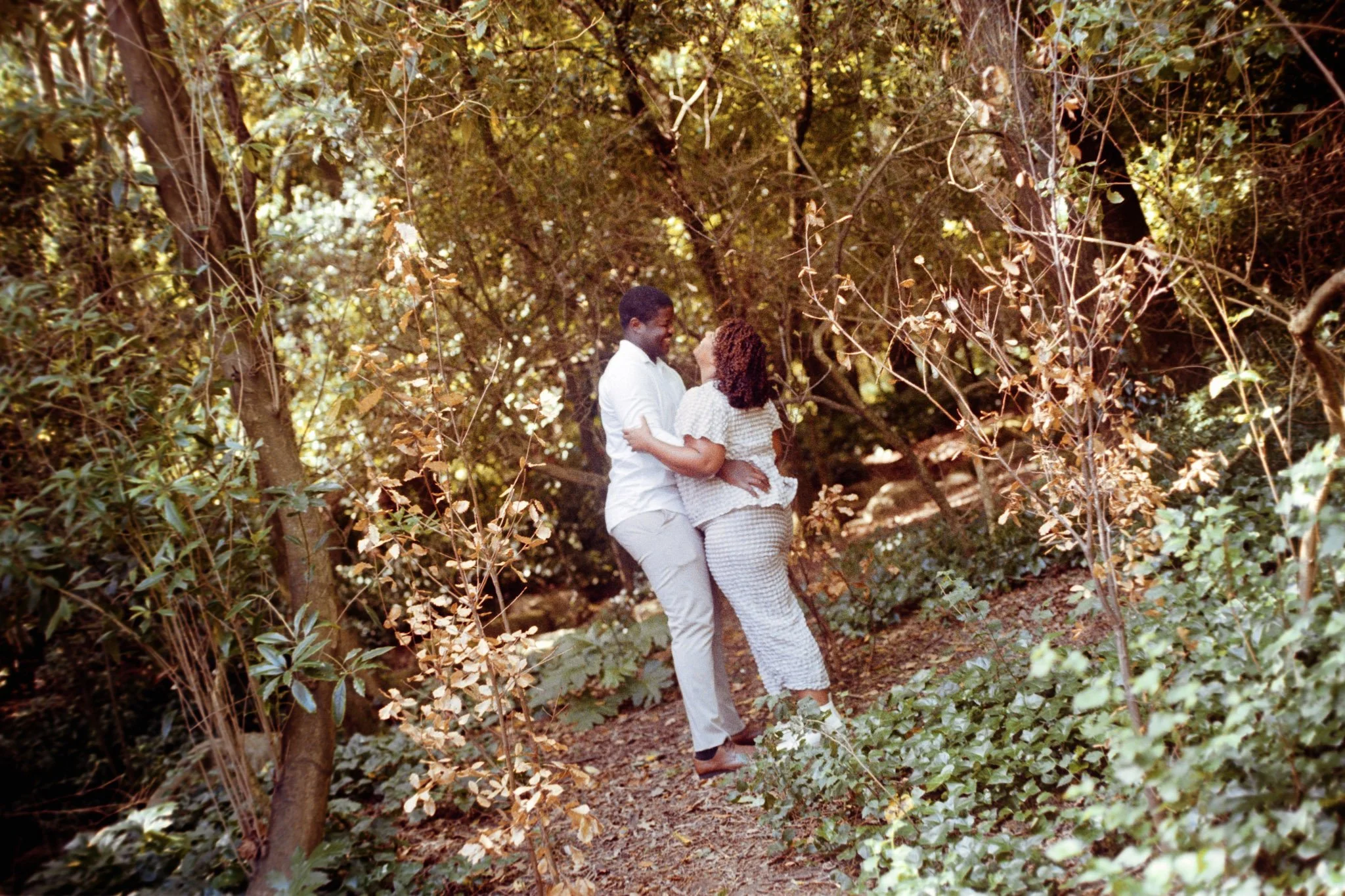 Casal dançando no meio de uma floresta densa com árvores e folhas ao redor.