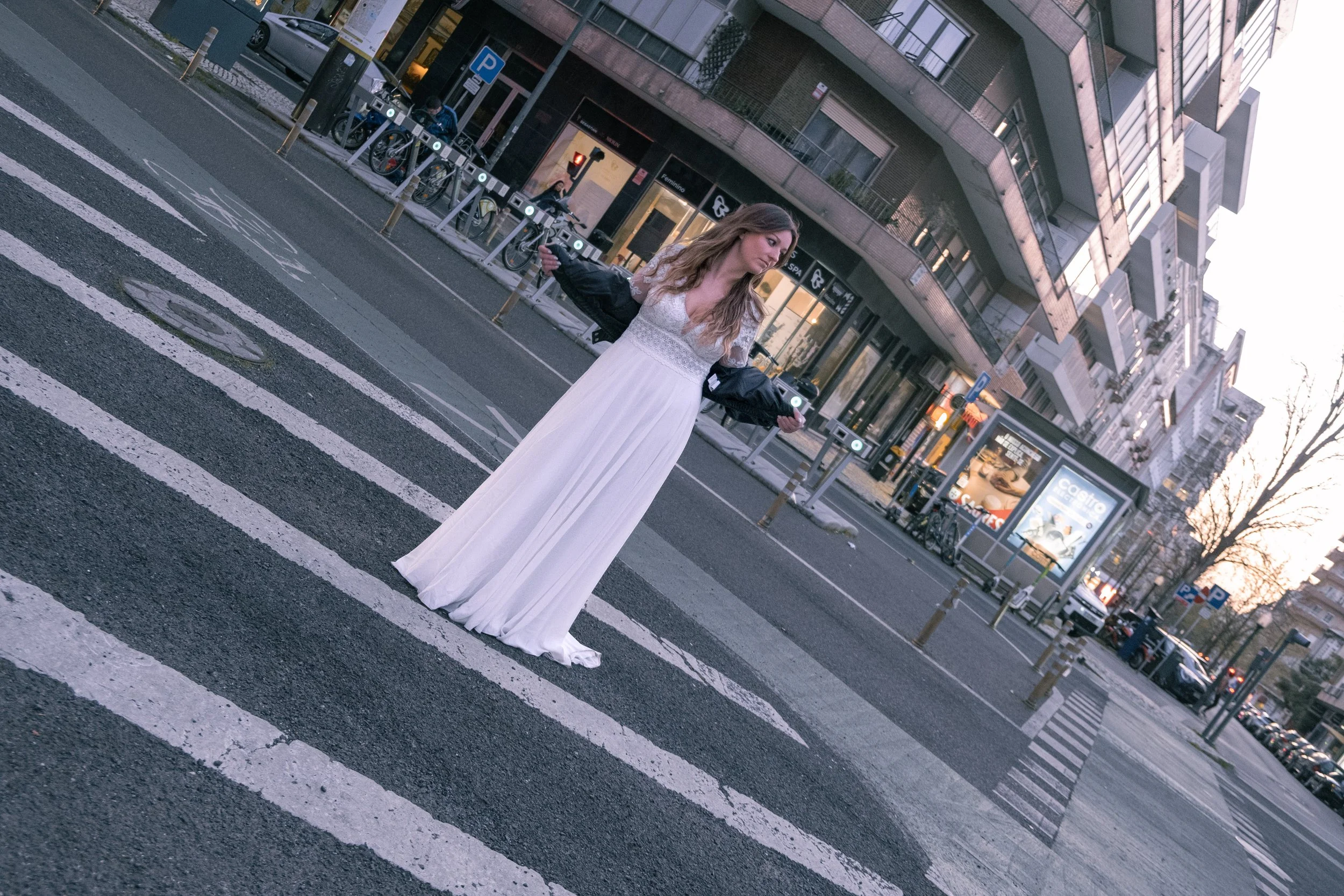 Pessoa de vestido branco em cruzamento de rua, segurando uma jaqueta preta, ao entardecer em uma área urbana com edifícios modernos ao fundo, bicicletas estacionadas e sinal de trânsito vermelho.