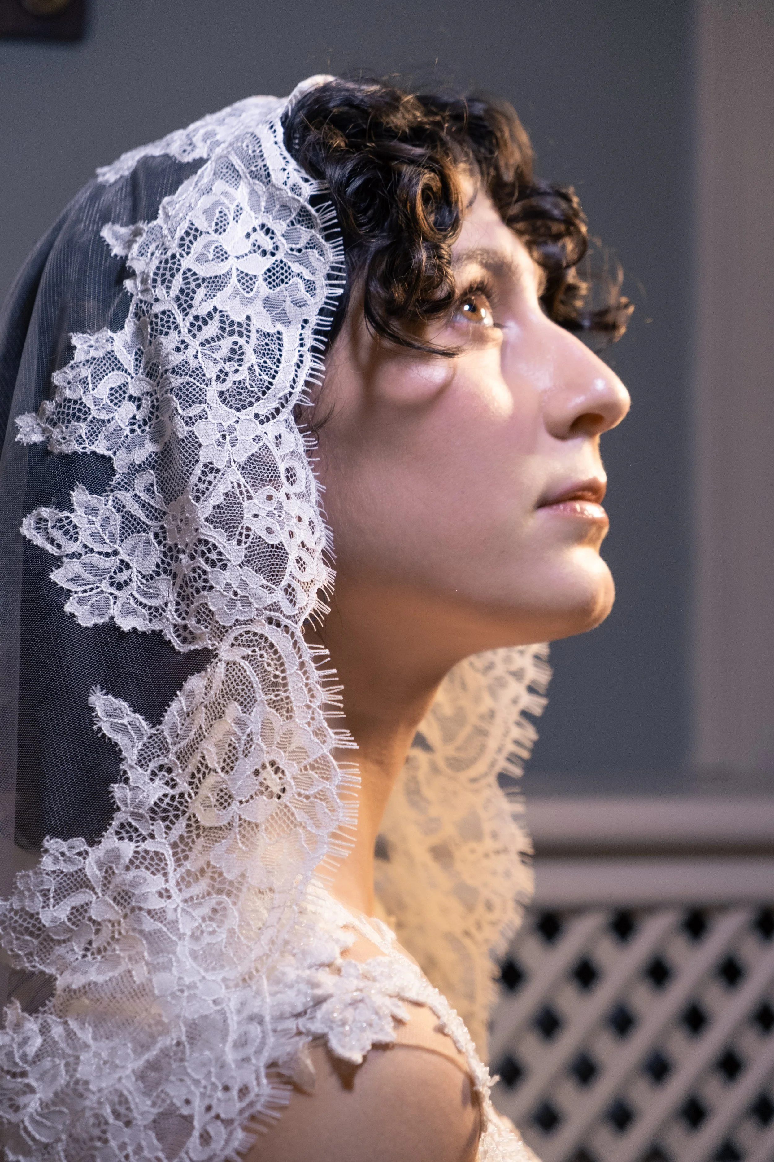 Retrato de uma mulher com cabelo cacheado, usando uma mantilha de renda branca, olhando para a direita, com fundo escuro.