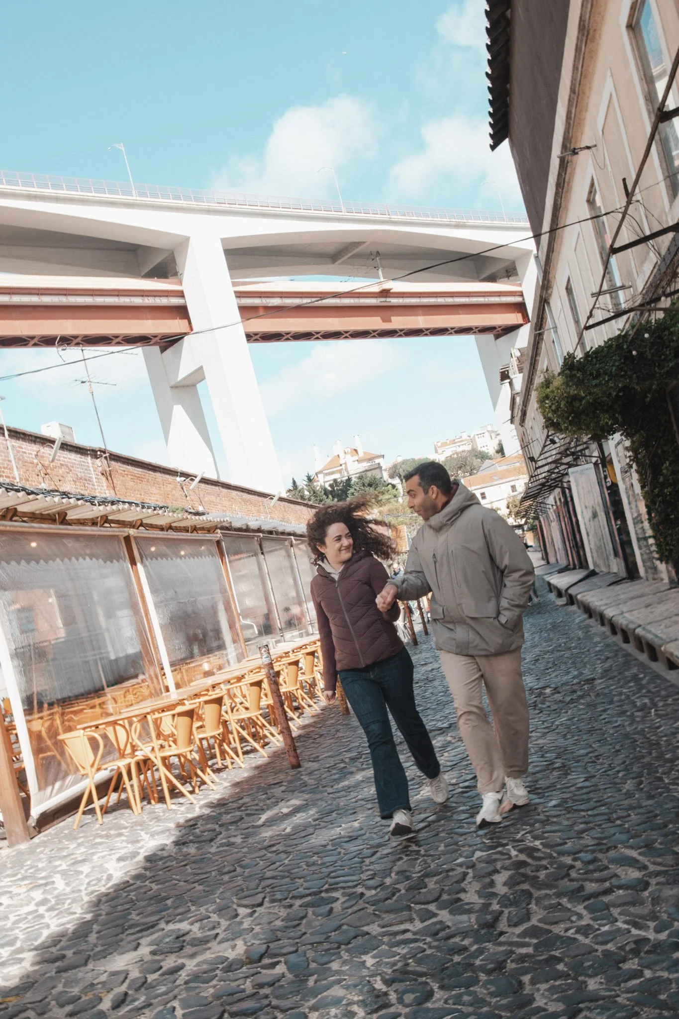 Homens conversando e caminhando na rua de pedra numa cidade com prédio de fachada antiga e um viaduto moderno no fundo.