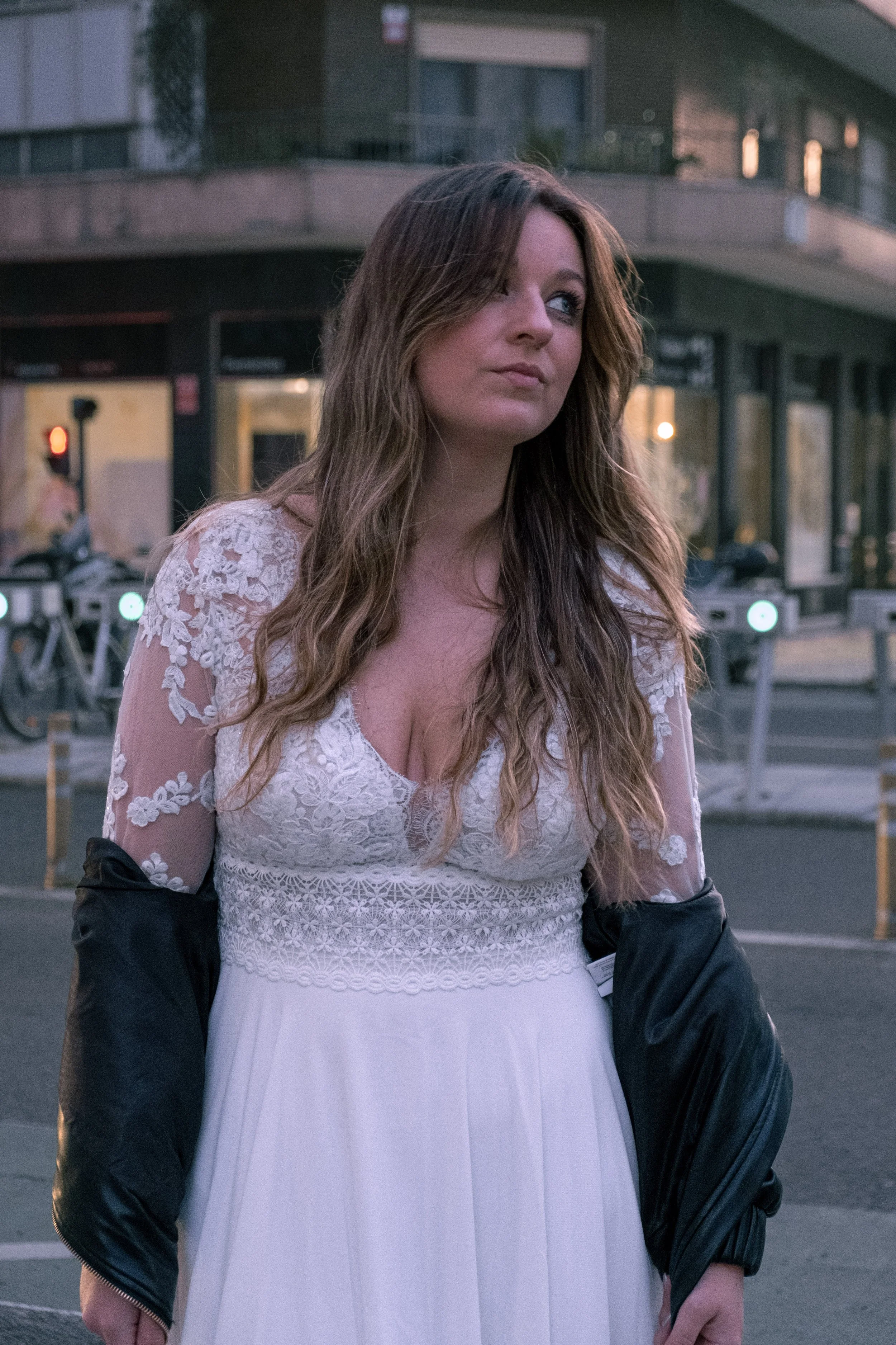 Mulher com cabelo castanho, usando vestido branco com detalhes em renda, e jaqueta preta. Ela está na rua, com fundo de prédio e alguns obstáculos de trânsito.