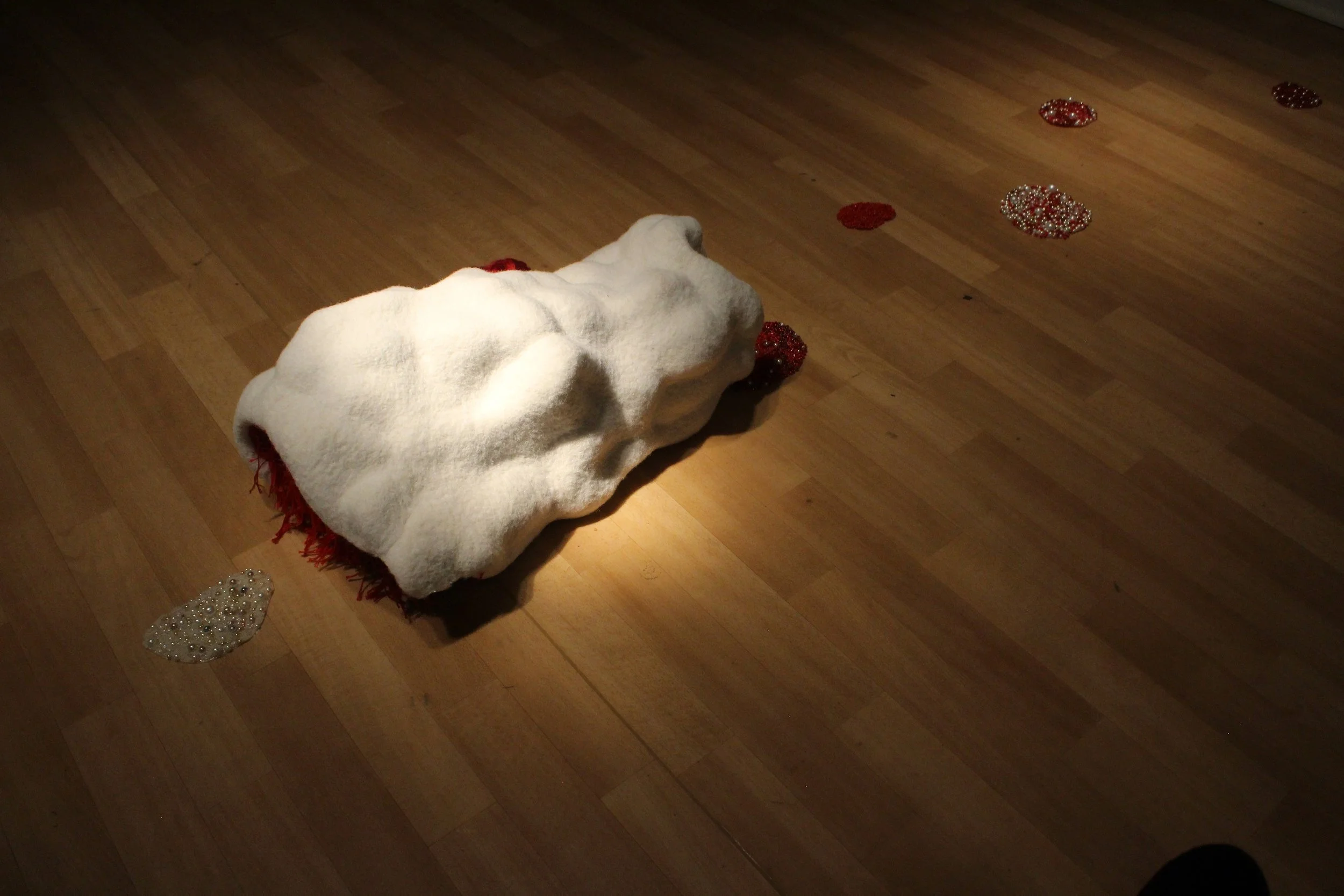 A white, shapeless, white fiber sculpture that lies on a hardwood floor surrounded by scattered red and silver decorative pearls.