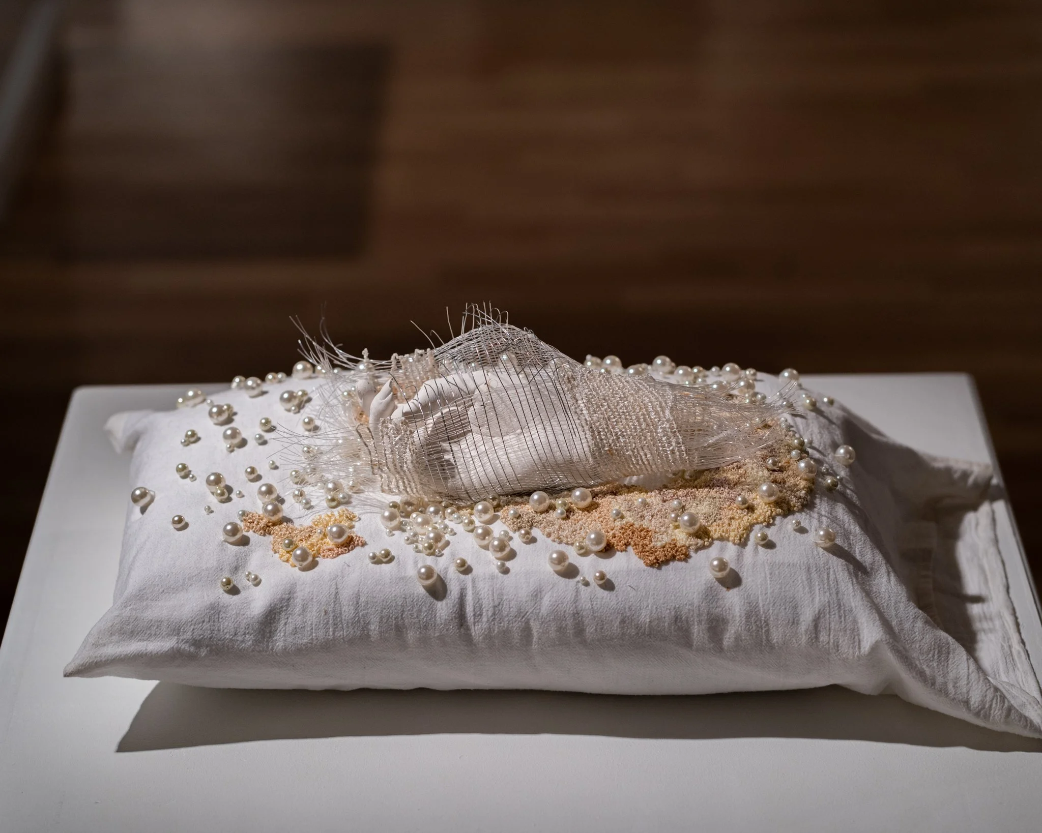 A white cushion decorated with pearl beads and intricate woven embellishments, displayed on a pillow with a plain cover.