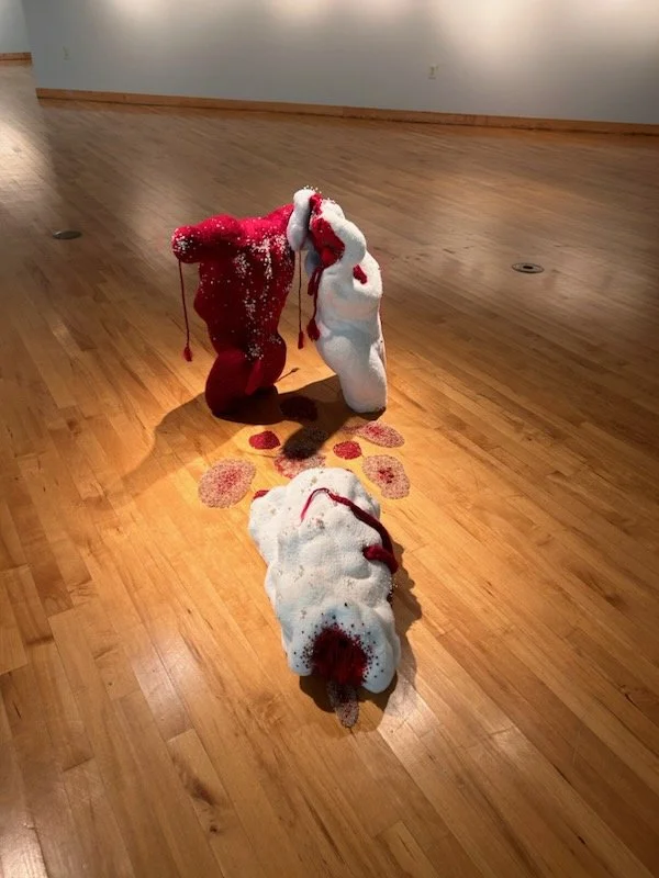 A white and red, torso sculpture that lies on a hardwood floor surrounded by scattered red and silver decorative pearls.