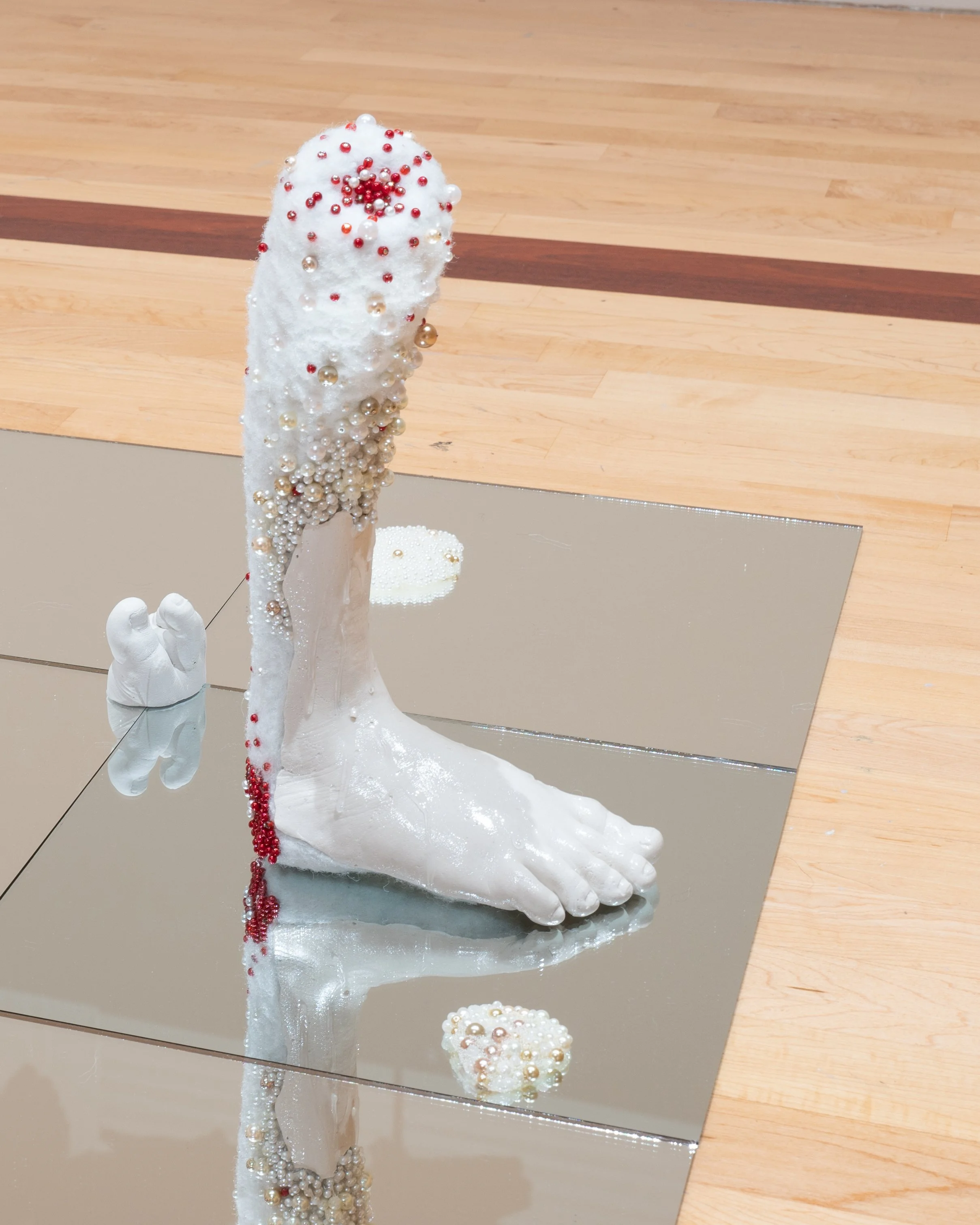 A white leg sculpture decorated with red, white, and gold beads, situated on a reflective surface with a wooden floor background.