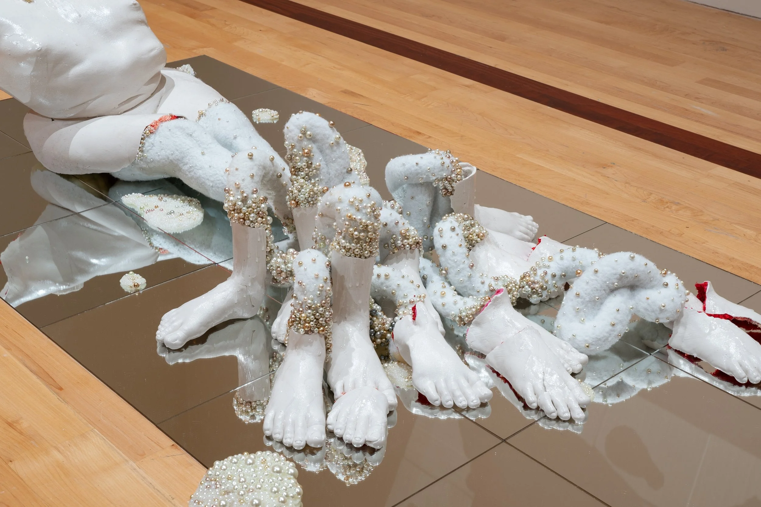 A sculpture of a reclining human figure with pearl embellishments, placed on a mirror surface with a wooden floor background.