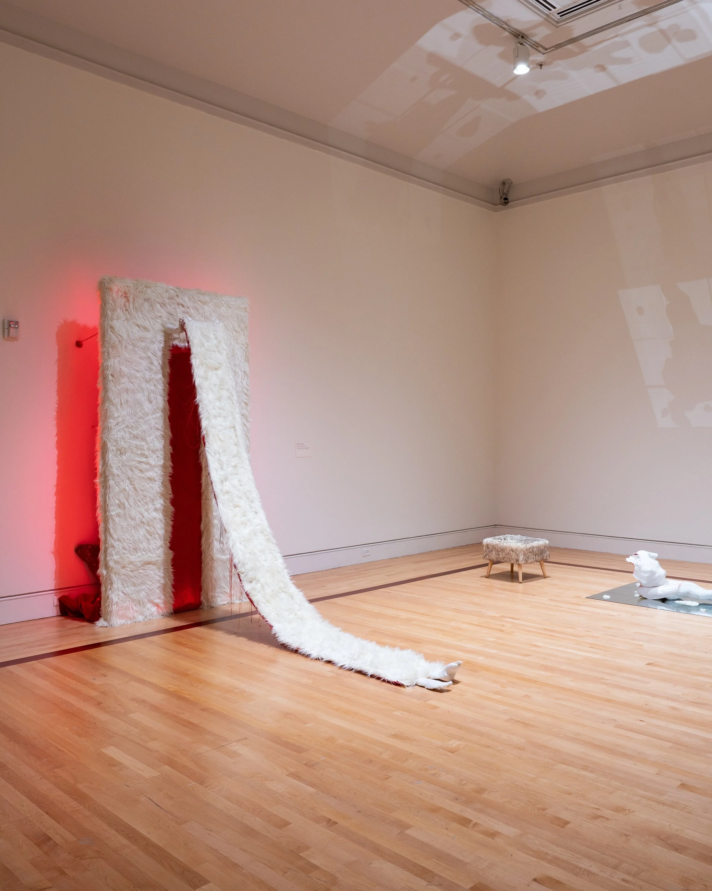 An art installation with a tall, plush, white fur-covered mirror frame on the floor, a small furry stool, and sculptures of a white animal, all in a gallery with minimal decor and hardwood floors.