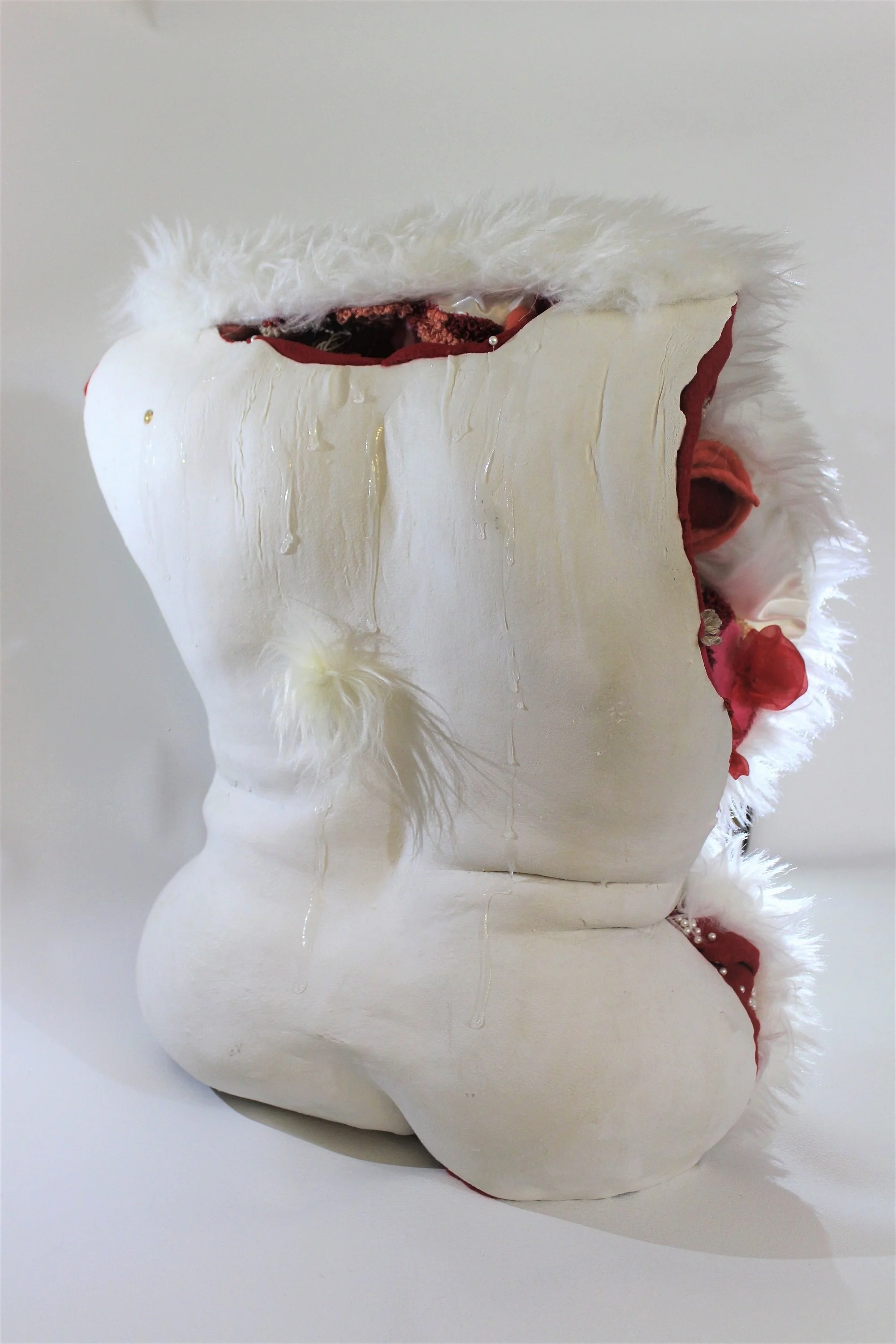 A white ceramic sculpture of a human torso decorated with red fabric, white faux fur trim, and pearls, with some liquid dripping down the surface.